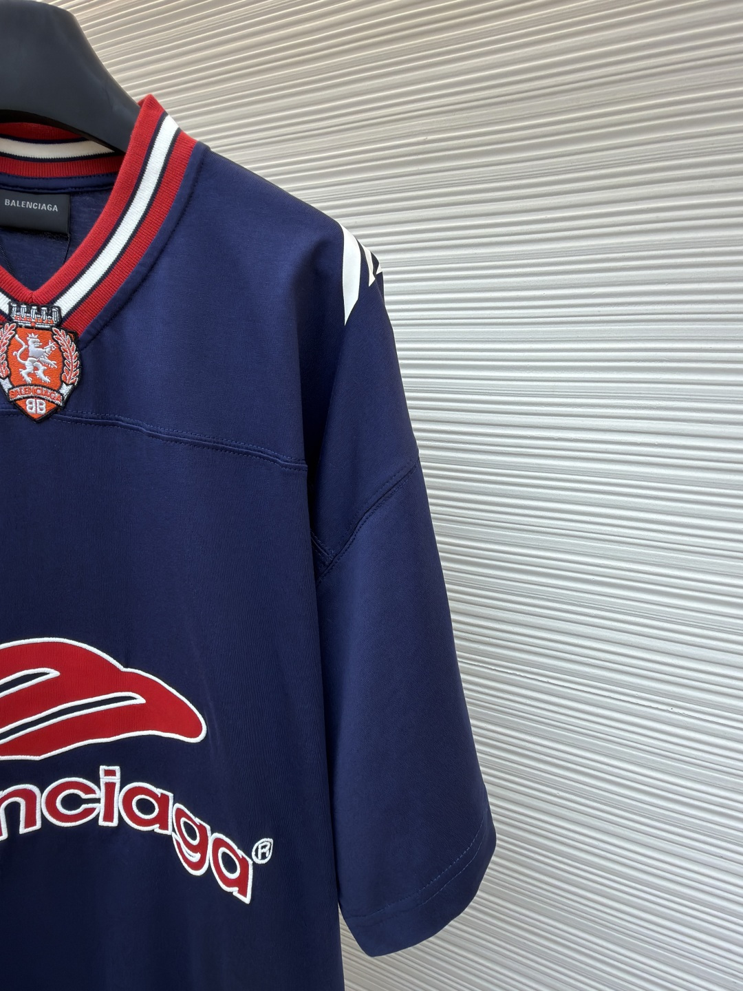 P180 Balenciaga 球队系列V领长袖
●原版：5699购入开发
●货号：1682041-A50
●面料：平纹针织紧密棉面料
●工艺：胶浆印花制作
●颜色：蓝色
●版型：宽松版型
●克重：约300克
●尺码：XS-S-M-L
●描述：主体采用24支特纺紧密棉，克重为185g/㎡。在纺纱环节应用了紧密纺技术，显著减少了纱线毛羽，使得纱线更光滑、强韧。这赋予了面料结构异常密实、表面光洁平整、抗起球性优异的特点，同时保留了纯棉的柔软透气性。185g的克重属于轻至中量级，确保了衣物轻盈、垂坠且活动自如的特性，非常适合打造兼具运动感与日常舒适度的长袖衫。V领部分由两种不同颜色的高弹罗纹面料拼接而成。其工艺步骤是先分别织造出两种颜色的罗纹带，再通过精准的剪裁与拼接，形成具有鲜明色彩对比的V形领口。打破了纯色衣身的单调，以强烈的色彩对比塑造出年轻、活力的视觉焦点，并优化了颈部线。将图案在不同材质或颜色的布料上预先裁剪成型贴布，然后将其覆盖在衣身上，最后用高密度绣线锁边固定并勾勒内部细节。其作用在于：创造色彩鲜明、质感对比强烈且极具立体感和耐用性的图案，图案边缘牢固，不易磨损。将特制的弹性浆料通过网版印刷在袖部的工艺，色彩鲜艳且具有轻微涂层感的图案。弹力浆料保证了印花区域能随面料一起拉伸而不开裂。与贴布绣的立体感形成一平一凸的质感对比，增加了设计的趣味性和复杂度。整体走线平直细腻，面料舒适柔软，颜色一致无色差 能与同款搭配营造时尚造型，驾驭各种场合，定制辅料，三标齐全，男女同款！