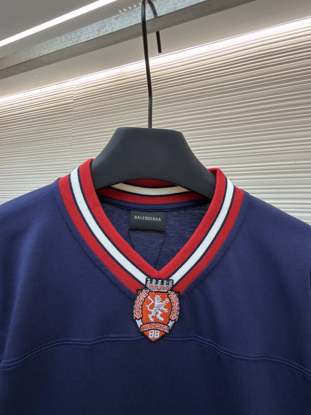 P180 Balenciaga 球队系列V领长袖
●原版：5699购入开发
●货号：1682041-A50
●面料：平纹针织紧密棉面料
●工艺：胶浆印花制作
●颜色：蓝色
●版型：宽松版型
●克重：约300克
●尺码：XS-S-M-L
●描述：主体采用24支特纺紧密棉，克重为185g/㎡。在纺纱环节应用了紧密纺技术，显著减少了纱线毛羽，使得纱线更光滑、强韧。这赋予了面料结构异常密实、表面光洁平整、抗起球性优异的特点，同时保留了纯棉的柔软透气性。185g的克重属于轻至中量级，确保了衣物轻盈、垂坠且活动自如的特性，非常适合打造兼具运动感与日常舒适度的长袖衫。V领部分由两种不同颜色的高弹罗纹面料拼接而成。其工艺步骤是先分别织造出两种颜色的罗纹带，再通过精准的剪裁与拼接，形成具有鲜明色彩对比的V形领口。打破了纯色衣身的单调，以强烈的色彩对比塑造出年轻、活力的视觉焦点，并优化了颈部线。将图案在不同材质或颜色的布料上预先裁剪成型贴布，然后将其覆盖在衣身上，最后用高密度绣线锁边固定并勾勒内部细节。其作用在于：创造色彩鲜明、质感对比强烈且极具立体感和耐用性的图案，图案边缘牢固，不易磨损。将特制的弹性浆料通过网版印刷在袖部的工艺，色彩鲜艳且具有轻微涂层感的图案。弹力浆料保证了印花区域能随面料一起拉伸而不开裂。与贴布绣的立体感形成一平一凸的质感对比，增加了设计的趣味性和复杂度。整体走线平直细腻，面料舒适柔软，颜色一致无色差 能与同款搭配营造时尚造型，驾驭各种场合，定制辅料，三标齐全，男女同款！
