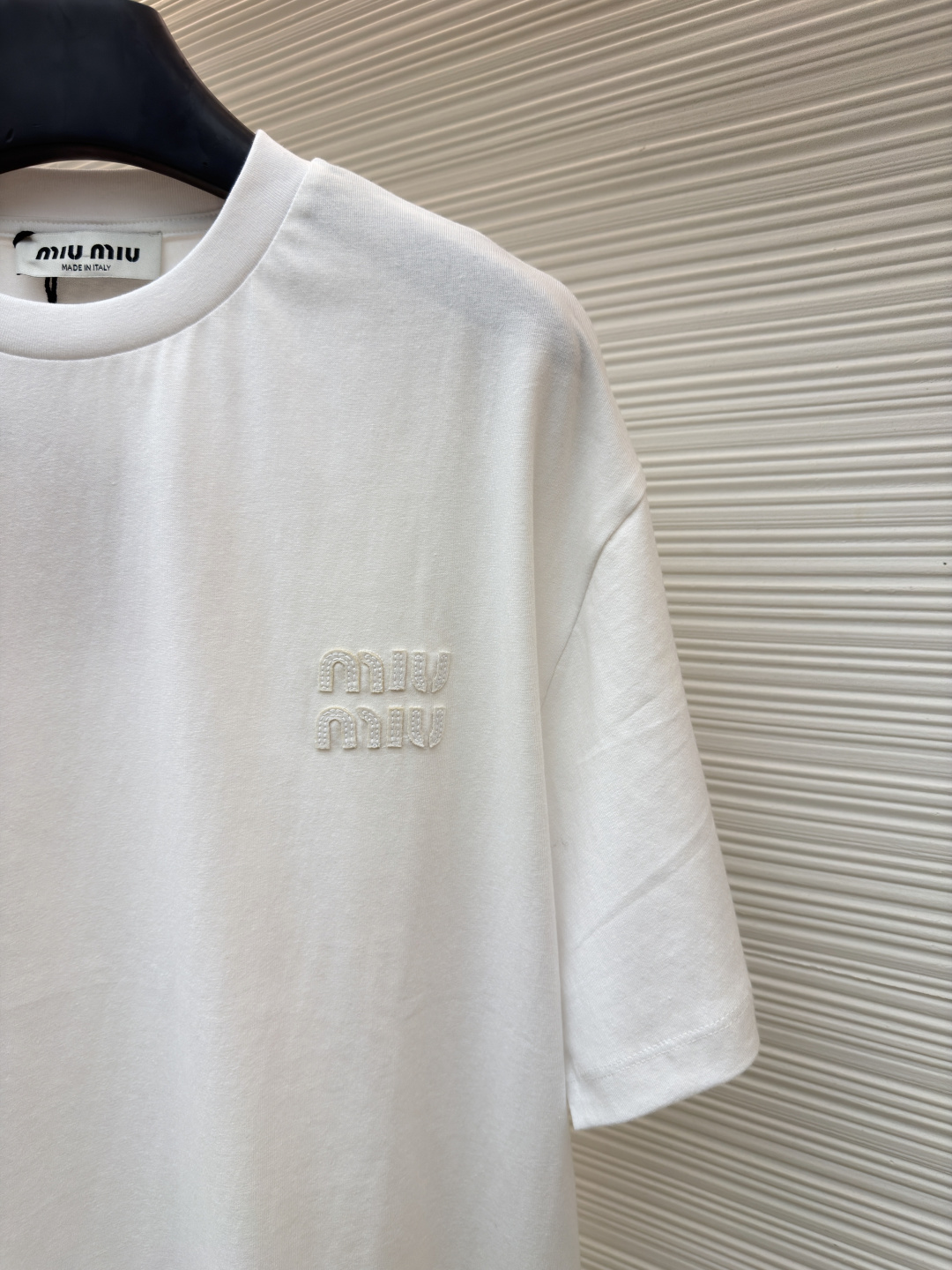 P125 MIU MIU经典字母logo图案短袖
●原版：5350购入开发
●货号：1682626-A00
●面料：精梳棉面料
●工艺：刺绣制作
●颜色：白色
●版型：合身版型
●克重：约220克
●尺码：S-M-L-XL
●描述：这款MIU MIU字母LOGO图案短袖以高支数精梳棉为面料基底，通过酵素洗水工艺处理，使布面触感更柔软顺滑，同时提升了面料的抗皱性与透气性，减少穿着紧绷感。胸前的品牌字母LOGO采用进口刺绣工艺，选用高光泽度的优质绣线，针脚细密均匀、图案立体饱满，兼具细腻质感与品牌辨识度，且经加固处理不易脱线。领口采用双针包边工艺，搭配同色系缝线，耐洗不易变形，贴合颈部曲线的同时保障了穿着舒适度。整体剪裁为宽松落肩版型，车线工整且线头处理干净，结合预缩水处理有效降低了洗涤后的缩水率，建议手洗以更好地保护刺绣细节与面料质感。整体走线平直细腻，面料舒适柔软，颜色一致无色差 能与同款搭配营造时尚造型，驾驭各种场合，定制辅料，三标齐全，男女同款！