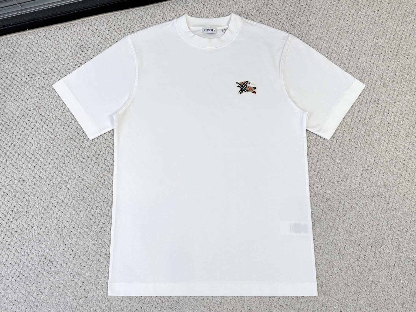 NO:833408,Product Introduction Burberry / Plaid logo cotton T-shirt Fabric 15 count special woven compact cotton / 230G Rib 32 count 1×1 rib / 340G  This short-sleeved piece is made of heavy-weight fabric with thick needles, which has strong texture and crisp texture. Since the 15-count yarn is relatively thick, the stiffness of the fabric supports the structure of the casual version. It can eliminate the internal stress generated during the weaving process of the thick-count yarn and prevent the shrinkage of the fabric caused by the subsequent embroidery process.  The pattern is misaligned. The neckline is made of pure cotton high-density tight ribbing, which achieves elasticity through high-density weaving and has a pure and natural touch. The overall fabric configuration provides a flat, stable and non-deformable base fabric for the tufted three-dimensional embroidery, and at the same time supports the structural sense of the casual version with a crisp texture.  The equestrian knight logo uses tufted three-dimensional toothbrush embroidery technology, which is an embroidery technique that uses special needles to embed embroidery threads into bundles into the base fabric to create an upright fluff effect. The embroidery needle pierces the fabric with high-speed reciprocating motion, bringing in multiple strands of embroidery thread at the same time and forming a loop; the embroidery is completed  Finally, the back side is coated with water-based adhesive to fix the bottom of the coil, and the front side is cut open and trimmed to a uniform height through a shearing process to form an upright texture similar to toothbrush bristle bundles.,,burberry,T-shirt,tees,t-shirt,19860909产品简介 巴宝莉 / 格纹徽标棉质 T恤 面料15支特纺紧密棉 / 230G 罗纹32支1×1罗纹 / 340G 这款短袖选用粗针重克重面料,肌理感强、质地挺括.由于15支纱线较粗,面料的挺括性支撑了休闲版型的结构感,它能消除粗支纱线织造过程中产生的内应力,防止后续刺绣工艺中因面料收缩导致图案错位.领口采用全棉高密度紧实罗纹,通过高密度织造实现弹性,触感纯粹自然.整体面料配置为簇绒立体刺绣提供了平整稳定、不易变形的底布基础,同时以挺括的质地支撑起休闲版型的结构感. 马术骑士徽标采用簇绒立体牙刷刺绣工艺,这是一种通过特殊针具将绣线成束植入底布、形成竖立状绒毛效果的刺绣技法.刺绣针以高速往复运动刺穿面料,将多股绣线同时带入并形成线圈；刺绣完成后,背面涂覆水性背胶固定线圈底部,正面则通过剪毛工序将线圈顶部剪开并修剪至统一高度,形成类似牙刷毛束的直立质感.,,burberry,T-shirt,tees，t-shirt,,Men's clothing