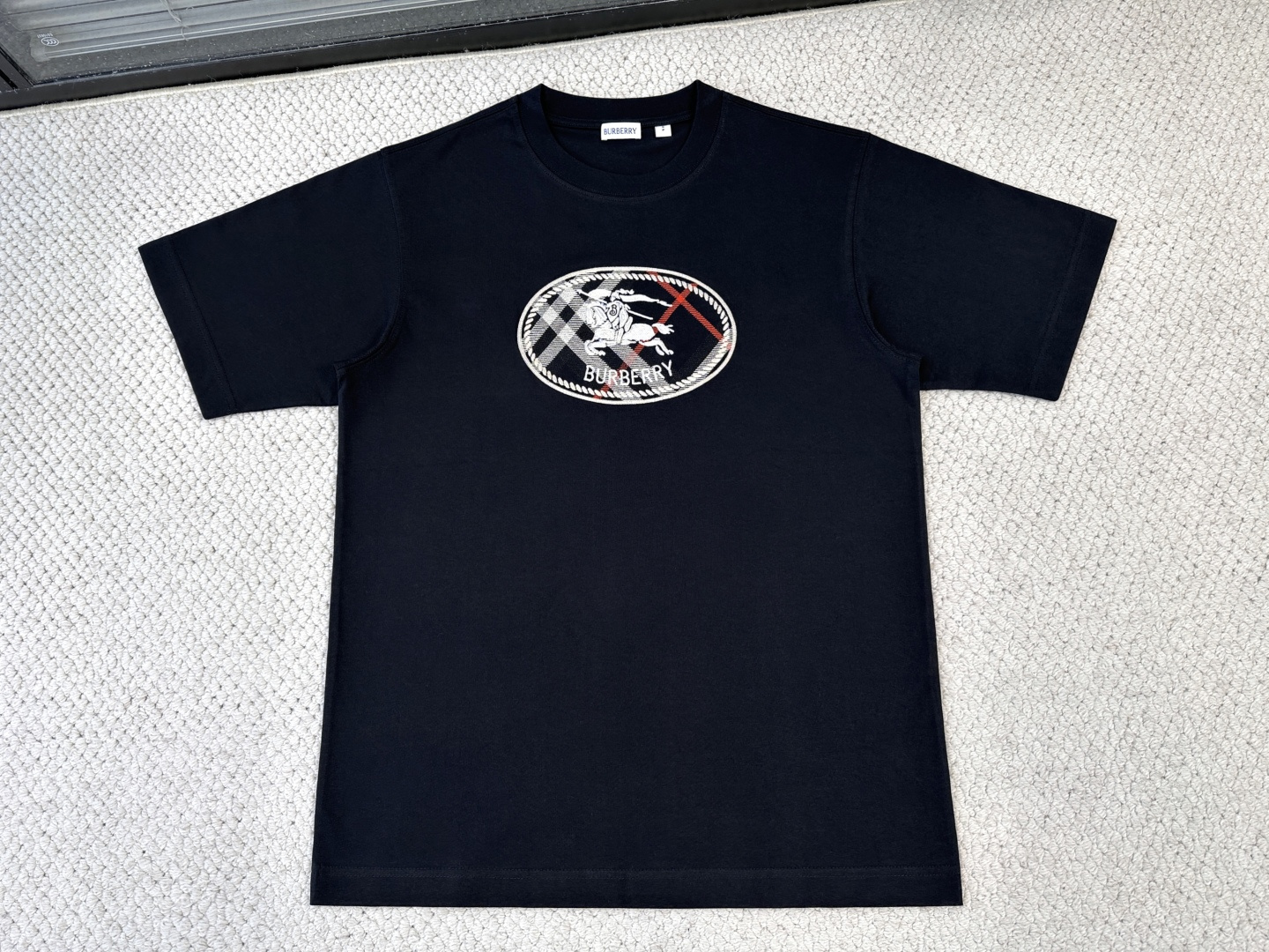 NO:833396,-Product Introduction Burberry / Plaid Knight Seal Cotton T-shirt Fabric 15-count special woven compact cotton / 230G rib with 32-count 1×1 rib / 340G This short-sleeved shirt is made of 15-count special-spun compact cotton, which is a heavy-gauge fabric with strong texture and crisp texture. Because the 15-count yarn is thicker, yarn hairiness is significantly reduced by improving the parallelism and cohesion of the fibers, giving the cloth a fine and slightly matte high-end texture, while greatly improving the anti-pilling level. The neckline is made of high-density cotton ribbing, which achieves elasticity through high-density weaving and has a pure and natural touch. The plaid knight's seal embroidery on the front chest uses a combination of multi-color chain embroidery and twill stitch embroidery. The chain embroidery is outlined along the outlines and color block boundaries of the plaid and knight's seal, creating rough and decorative line edges, with a vivid feel similar to hand-drawn graffiti. In the area framed by chain stitch, twill stitches are used to fill the whole area; the stitches are arranged at specific angles to produce a delicate glossy flow, simulating the visual effect of the twill texture of the fabric. The twill stitches have a stronger three-dimensional effect and anti-pilling properties, and can effectively avoid the problem of shrinkage and stiffness of the fabric caused by large-area embroidery.,,burberry,T-shirt,tees,t-shirt,19860909-产品简介 巴宝莉 / 格纹骑士印章棉质T恤衫 面料15支特纺紧密棉 / 230G 罗纹搭配32支1×1罗纹 / 340G 这款短袖选用15支特纺紧密棉,属于粗针重克重面料,肌理感强、质地挺括.由于15支纱线较粗,通过提高纤维的平行度和抱合力,显著减少了纱线毛羽,使得布面呈现出细腻且微雾面的高端质感,同时大幅提升了抗起毛球等级 .领口采用全棉高密度紧实罗纹,通过高密度织造实现弹性,触感纯粹自然. 前胸格纹骑士印章刺绣采用多色锁链绣搭配斜纹挨针刺绣的组合工艺.锁链绣沿格纹和骑士印章的轮廓及色块边界进行勾勒,出粗犷且富有装饰性的线条边缘,具有类似手绘涂鸦的生动感. 在锁链绣框定的区域内,采用斜纹走向的挨针满幅填充；针迹按照特定角度排列,产生细腻的光泽流动感,模拟出织物的斜纹肌理视觉效果.斜纹挨针的立体感和抗起毛性更强,且能有效避免大面积刺绣导致的面料收缩板硬问题.,,burberry,T-shirt,tees，t-shirt,,Men's clothing