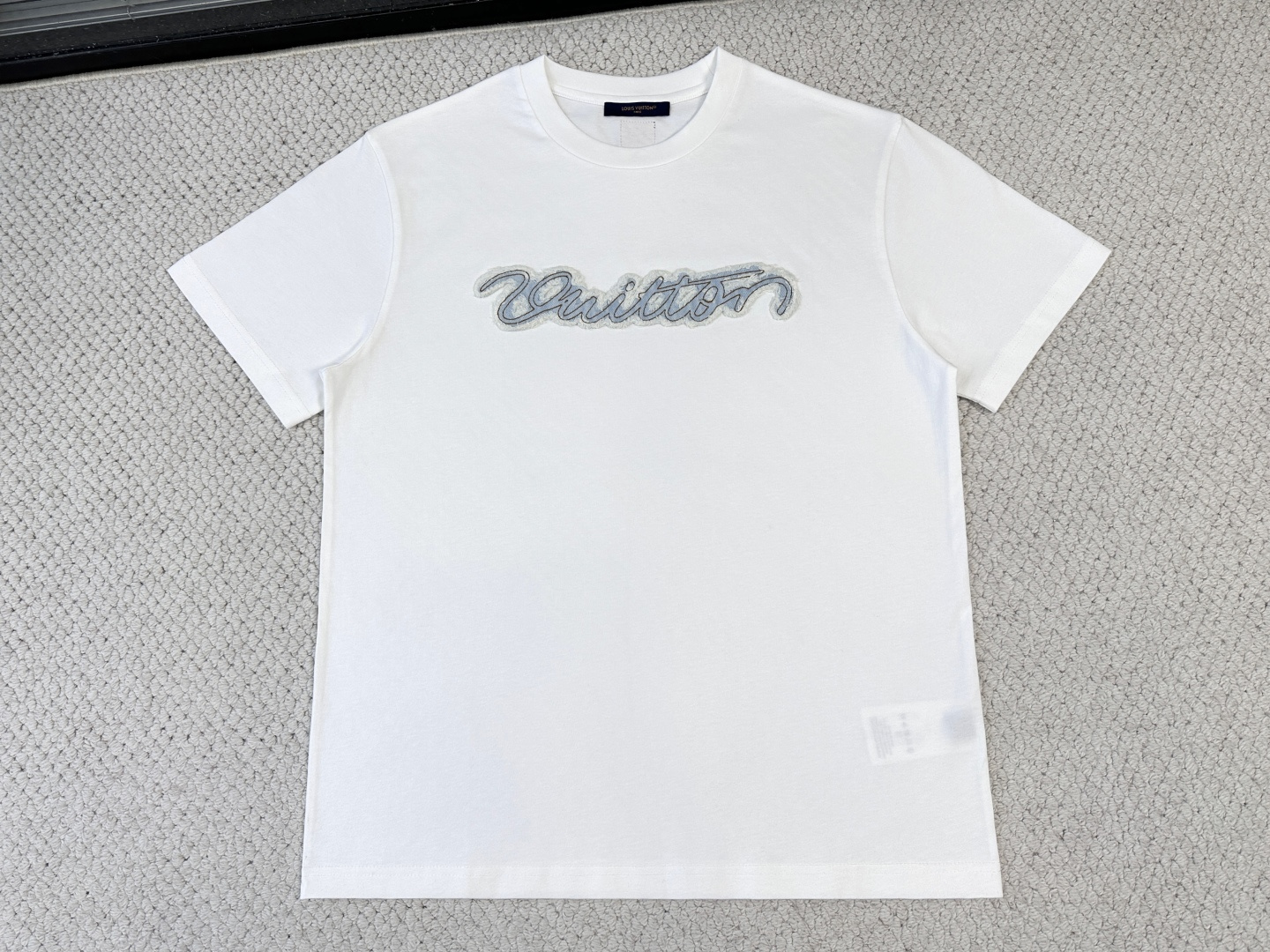 NO:840548,LV Product Introduction 1V / Logo Denim T-shirt Fabric: 15-count special woven compact cotton / 230G Rib: 32-count 1×1 rib / /340G This classic T-shirt is embellished with a handwritten denim patch on the chest, capturing the distressed effect and textured embroidery technology. The woven patch at the back adds subtle details. The casual design is easy to match and easily upgrades the summer casual look. This short-sleeved shirt is made of 15-count special-spun compact cotton. The thicker yarn gives the fabric obvious texture and stiffness. Mercerizing treatment makes the cross-section of the cotton fiber round, significantly enhances the gloss and uniformity of dye adsorption, and makes the blue and white background colors fuller and more uniform. The ribbed neckline retracts to its optimal elastic state during the shaping process, and the compact structure is fully stable, forming a collar that fits well without being pinched. The patch itself is made of denim fabric, which has been precision laser cut into the shape of handwritten letters. Before sewing, it is independently washed and aged, and pumice and low-concentration bleach are added to produce irregular color fading, whitening, and slight yarn breakage on the surface, simulating the natural fading and wear effects after years of wearing. The positioning embroidery process is carried out with precision routing along the edge of the patch, and the stitches are closely arranged, which not only plays a fixing role and prevents the edge from falling off after washing, but also simulates the visual effect of handwriting routing. The rear woven label is woven by a jacquard loom, and the edges are thermally cut to prevent stripping. It is fixed with four-corner topstitching, forming a level of detail that echoes the front and rear chest patches.,, louis vuitton, T-shirt, tees, t-shirt,19860909LV 产品简介 1V / 标识牛仔T恤 面料15支特纺紧密棉 / 230G 罗纹32支1×1罗纹 / /340G 本款经典T恤于胸前点缀手写体牛仔贴饰,撷取做旧效果纹理感刺绣工艺呈现.后部梭织贴饰平添微妙细节.随性设计易于搭配,轻松升格夏日休闲造型. 这款短袖选用15支特纺紧密棉,较粗纱线赋予织物明显的肌理感与挺括度.丝光处理使棉纤维截面圆润,显著增强光泽度与染料吸附均匀性,使蓝色与白色的底色更加饱满匀净.罗纹的领口在定型过程中回缩至最佳弹性状态,紧实结构充分稳定,形成贴合而不勒的领型. 贴饰本身为牛仔布料,经精密激光切割裁切成手写体字母形态；缝制前独立经历洗水做旧处理,加入浮石与低浓度漂白剂,表面产生不规则的色落、磨白和轻微的纱线断裂,模拟经年穿着后的自然褪色与磨损效果. 定位刺绣工艺沿贴饰边缘进行精密走线,针迹紧密排列,既起到固定作用、防止洗涤后翘边脱落,又模拟了手写体走线的视觉效果.后部梭织贴标由提花梭织机织出,边缘经热切割防脱丝,采用四角明线固定,与前胸贴饰形成前后呼应的细节层次.,,louis vuitton,T-shirt,tees，t-shirt,,Men's clothing