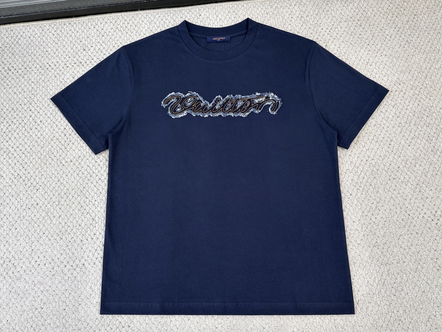 NO:840554,Lv logo denim T-shirt. Fabric: 15-count special woven compact cotton / 230G. Rib: 32-count 1×1 rib / /340G. This classic T-shirt is embellished with a handwritten denim patch on the chest, capturing the distressed effect and textured embroidery technology. The woven patch on the back adds subtle details. The casual design is easy to match, and it can easily upgrade the summer casual look. This short-sleeved shirt is made of 15-count special-spun compact cotton. The thicker yarn gives the fabric obvious texture and stiffness. Mercerizing treatment makes the cross-section of the cotton fiber round, significantly enhances the gloss and uniformity of dye adsorption, and makes the blue and white background colors fuller and more uniform. The ribbed neckline retracts to its optimal elastic state during the shaping process, and the compact structure is fully stable, forming a collar that fits well without being pinched. The patch itself is made of denim fabric, which has been precision laser cut into the shape of handwritten letters. Before sewing, it is independently washed and aged, and pumice and low-concentration bleach are added to produce irregular color fading, whitening, and slight yarn breakage on the surface, simulating the natural fading and wear effects after years of wearing. The positioning embroidery process is carried out with precision routing along the edge of the patch, and the stitches are closely arranged, which not only plays a fixing role and prevents the edge from falling off after washing, but also simulates the visual effect of handwriting routing. The rear woven label is woven by a jacquard loom, and the edges are thermally cut to prevent stripping. It is fixed with four-corner topstitching, forming a level of detail that echoes the front and rear chest patches.,, louis vuitton, T-shirt, tees, t-shirt,19860909Lv 标识牛仔T恤 面料15支特纺紧密棉 / 230G 罗纹32支1×1罗纹 / /340G 本款经典T恤于胸前点缀手写体牛仔贴饰,撷取做旧效果纹理感刺绣工艺呈现.后部梭织贴饰平添微妙细节.随性设计易于搭配,轻松升格夏日休闲造型. 这款短袖选用15支特纺紧密棉,较粗纱线赋予织物明显的肌理感与挺括度.丝光处理使棉纤维截面圆润,显著增强光泽度与染料吸附均匀性,使蓝色与白色的底色更加饱满匀净.罗纹的领口在定型过程中回缩至最佳弹性状态,紧实结构充分稳定,形成贴合而不勒的领型. 贴饰本身为牛仔布料,经精密激光切割裁切成手写体字母形态；缝制前独立经历洗水做旧处理,加入浮石与低浓度漂白剂,表面产生不规则的色落、磨白和轻微的纱线断裂,模拟经年穿着后的自然褪色与磨损效果. 定位刺绣工艺沿贴饰边缘进行精密走线,针迹紧密排列,既起到固定作用、防止洗涤后翘边脱落,又模拟了手写体走线的视觉效果.后部梭织贴标由提花梭织机织出,边缘经热切割防脱丝,采用四角明线固定,与前胸贴饰形成前后呼应的细节层次.,,louis vuitton,T-shirt,tees，t-shirt,,Men's clothing