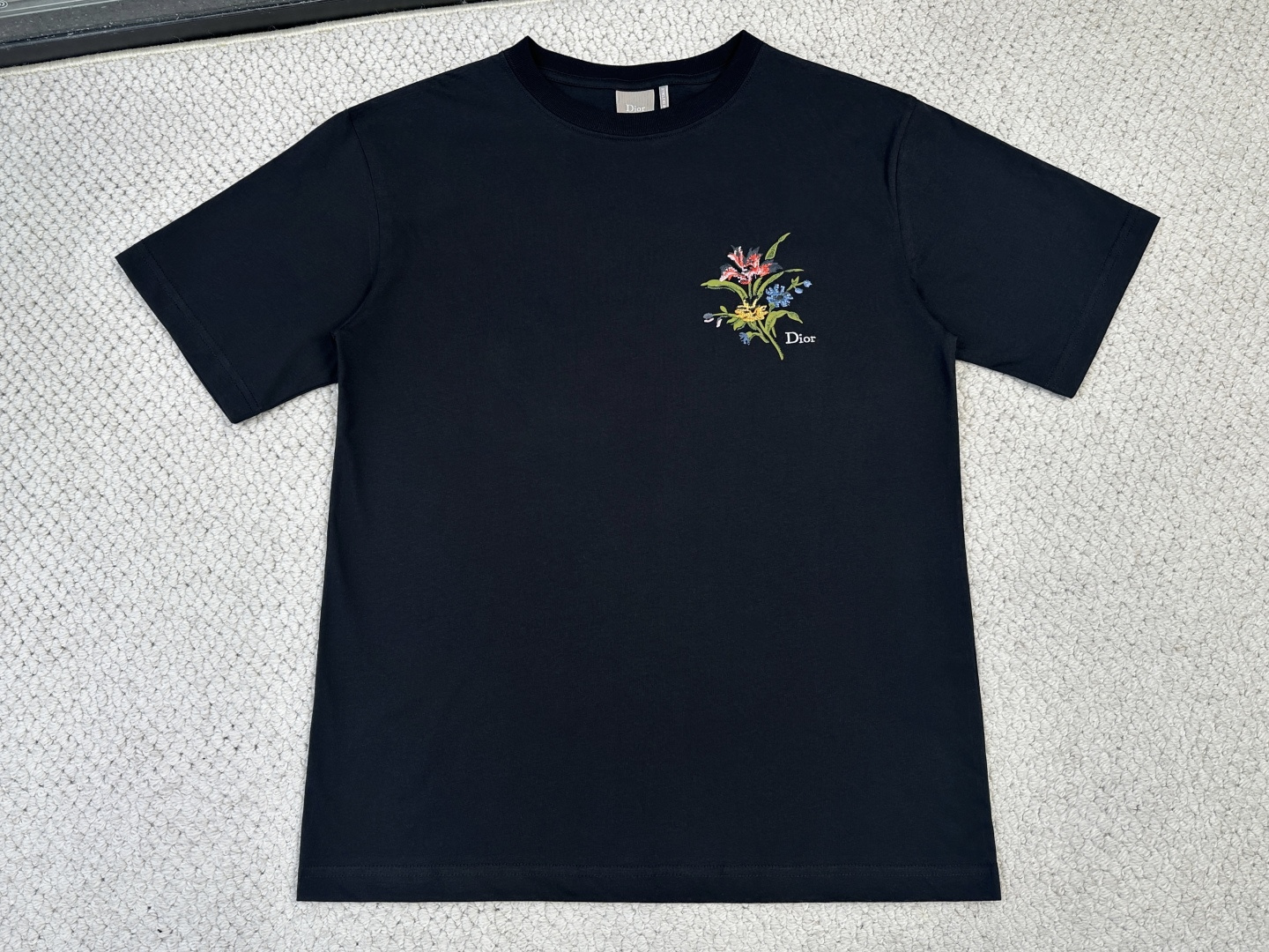 NO:840542,Dior - Product Introduction CD / Secret Garden graphic T-shirt Fabric 24-count double-sided plain cotton fabric / 250G Rib 24-count 1×1 rib / 200G This T-shirt pays tribute to the exquisite craftsmanship of Dior. It is carefully made of white cotton jersey fabric and decorated with the Secret Garden pattern, inspired by the classic floral tapestry, exuding a poetic and delicate atmosphere. It can be matched with the shorts of this series.  Count 24 is a medium-to-high fine yarn count. The yarn is fine, and the cloth surface is delicate and smooth, with a sense of refinement similar to that of a high-end shirt. The double-sided knitted structure has a fluffy and soft feel and drape support, and the upper body is not soft. The surface of the fabric presents an elegant slightly matte texture, which is smooth but not cold to the touch. It has a stiff frame and is skin-friendly and breathable. The rib yarn count is consistent with the main fabric, and the weight is slightly lower, forming a naturally fitting neckline trim.  The bouquet pattern adopts a composite process of penetration printing and positioning pin bionic embroidery. Penetration printing uses special additives to completely penetrate the dye molecules into the cotton fiber, restoring the rich color gradients and delicate brushstroke levels to the greatest extent. The thickness and softness of the fabric in the pattern area and the blank area are exactly the same, maintaining the original silky touch of double-sided cotton.  The positioning needle bionic embroidery positioning mark accurately embroiders each stitch in the corresponding area. The embroidery thread forms a coil on the back of the fabric, and the front shows a fluffy yarn arrangement, which has a natural texture similar to floral fluff and plant fiber. The embroidery is only selectively embellished on the outline of the flowers, so that the embroidery forms a three-dimensional decorative effect of blending of printing and embroidery from the printing.,, dior, shorts, shirts, tees, t-shirt,19860909迪奥-产品简介 CD / Secret Garden 图案T恤 面料24支双面平纹纯棉面料 / 250G 罗纹24支1×1罗纹 / 200G 这款T恤向的精湛工艺致敬.采用白色棉质平纹针织面料精心制作,饰以Secret Garden 图案,灵感源自经典花卉挂毯,散发诗意与精致的气息.可与该系列的短裤搭配. 24支属于中高细纱支,纱线较细,布面细腻光滑,具有近似高级衬衫的精致感.双面针织结构兼具蓬松柔软的手感与垂坠支撑力,上身不软塌.面料表面呈现优雅的微哑光质感,触感柔滑而不冰冷,挺括骨架与亲肤透气并存.罗纹纱支与主布一致,克重略低,形成自然贴合的领口饰边. 花束图案采用渗透印花融入定位插针仿生刺绣的复合工艺.渗透印花通过特殊助剂使染料分子完全渗入棉纤维内部,最大程度还原丰富的色彩渐变与细腻笔触层次,图案区域与空白区域的面料厚度、柔软度完全一致,保持了双面棉原有的丝滑触感. 定位插针仿生刺绣定位标记,精确将每一针绣在对应区域,绣线在面料背面形成线圈,正面呈现蓬松的纱线排列,具有类似花卉绒毛和植物纤维的天然质感.刺绣仅选择性地点缀在花朵轮廓,使刺绣从印花中形成印绣交融的立体装饰效果.,,dior,shorts,shirts,tees，t-shirt,,Men's clothing