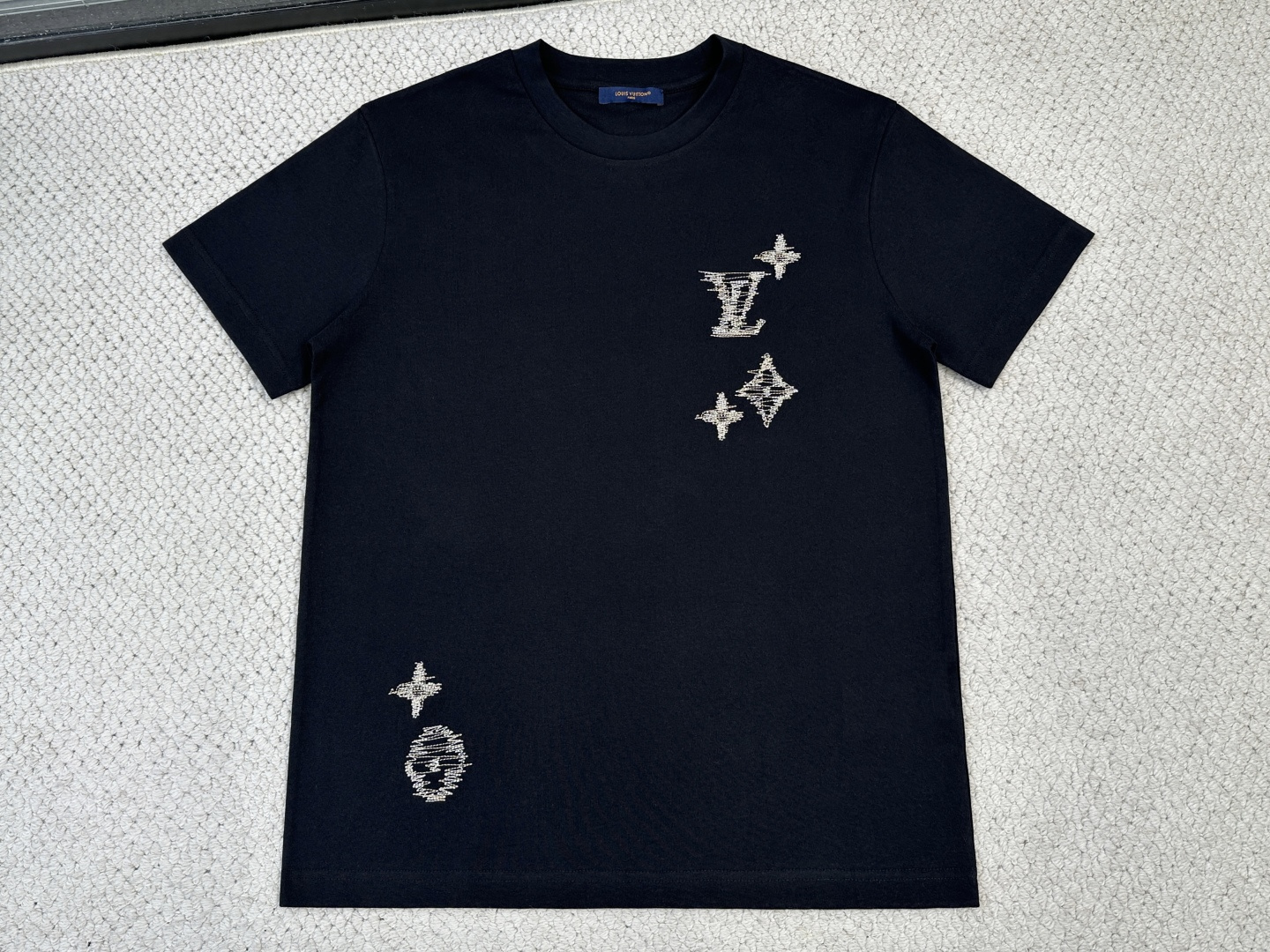 NO:843429,Lv Product Introduction 1V / Sashiko embroidered logo T-shirt Fabric 15-count special woven compact cotton plain fabric / 230G Rib 32-count 1×1 rib / 340G This lightweight summer cotton T-shirt has a soft texture and is decorated with the series' iconic Sashiko embroidery. The design is inspired by traditional Japanese craftsmanship, with textured logos embroidered with contrasting yarns. The precision workmanship injects ingenious style into the classic wardrobe. The short sleeves are made of 15-count special-spun compact cotton plain fabric. The special-spun compact spinning process significantly reduces yarn hairiness and has a high pilling resistance level. The medium-high weight gives the fabric excellent bone texture and drape, which is crisp but not stiff. The neckline is made of 32-count spun yarn with ultra-high weight ribbing. It is extremely dense and thick, has high-strength binding force, and can firmly lock the neck arc. The embroidery logos embellished on the front and back adopt Sashiko style craftsmanship, with textured logos embroidered with contrasting yarns. Typical Sashiko embroidery uses thickly twisted cotton thread, and the stitches present a vague undulating texture on the surface of the fabric. Thickly twisted cotton embroidery thread is used in obvious contrast with the background color of the fabric, and the surface of the embroidery thread is slightly hairy. The computer embroidery machine smoothes the curves by shortening the length of the stitches and prevents jagged edges at the turning points. The stitches are arranged in parallel to outline the outline, and the small gaps between the stitches allow the background color of the fabric to faintly show through, forming a unique texture and handmade texture.,,louis vuitton,T-shirt,tees,t-shirt,19860909Lv 产品简介 1V / Sashiko刺绣标识T 恤 面料15支特纺紧密棉平纹面料 / 230G 罗纹32支1×1罗纹 / 340G 本款轻盈夏日棉质T恤质感柔和,饰有本系列标志性Sashiko刺绣.设计灵感源自日本传统工艺,以撞色纱线绣饰纹理感标识.精工之作为经典衣橱注入匠心格调. 短袖选用15支特纺紧密棉平纹面料,特纺紧密纺工艺显著减少了纱线毛羽,同时抗起毛球等级高.中高克重赋予面料极佳的骨感与垂坠性,挺括而不僵硬.领口采用32支细纱配合超高克重罗纹,极其致密厚重,具有高强度收束力,能牢牢锁定颈部弧线. 前后点缀的刺绣标识采用刺子绣风格工艺,以撞色纱线绣饰纹理感标识.典型刺子绣使用粗捻棉线,线迹在面料表面呈现隐约的起伏质感.使用与面料底色形成明显对比的粗捻棉绣线,绣线表面略带毛羽. 电脑绣花机通过缩短针步长度来平滑曲线,防止转折处出现锯齿状.针迹以平行排列的方式勾勒轮廓,针迹间的微小间隙让面料底色隐约透出,形成独特的纹理感与手工质感.,,louis vuitton,T-shirt,tees，t-shirt,,Men's clothing