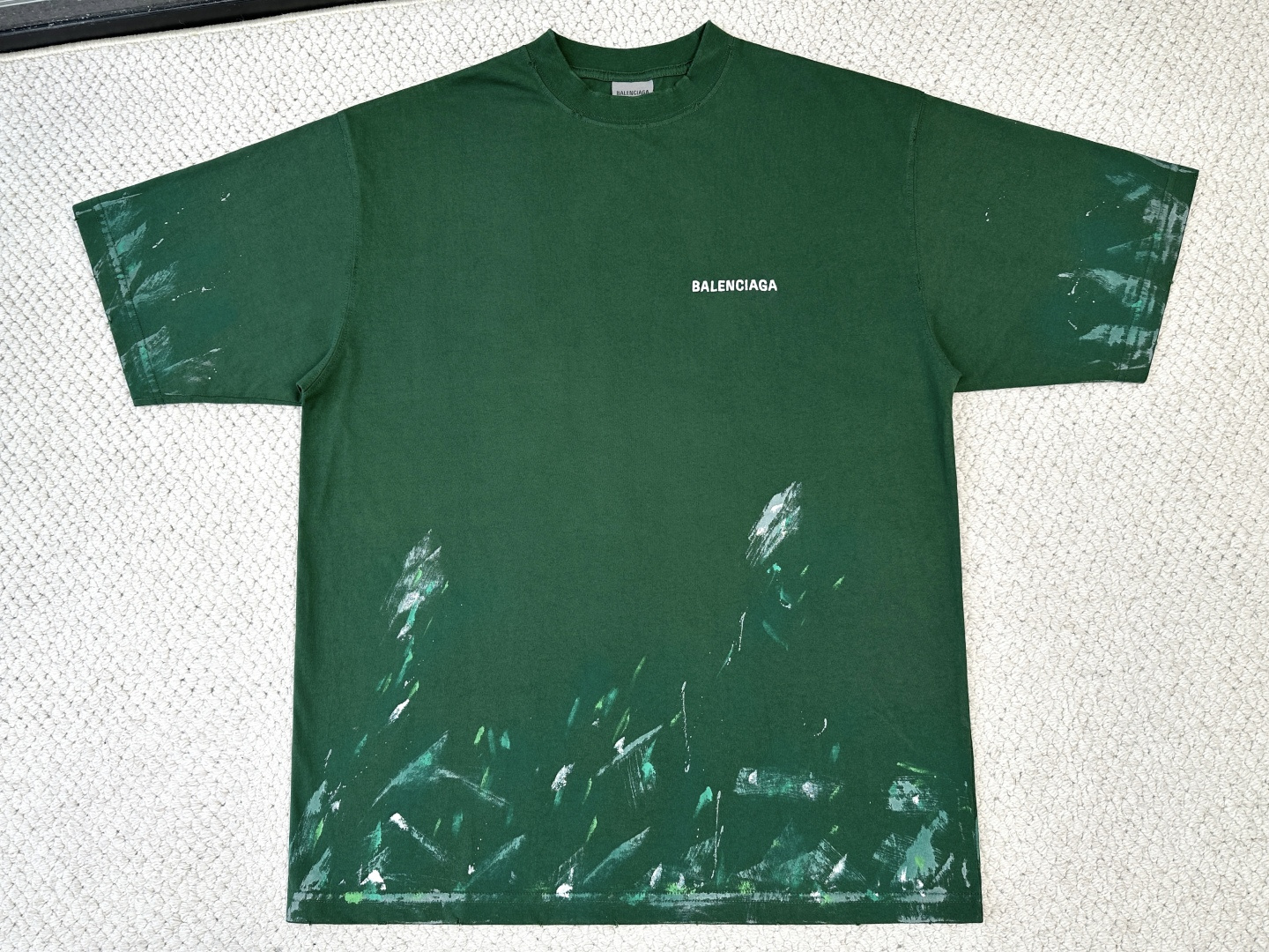 NO:914776,Balenciaga / Product Introduction Paris / Graffiti Loose Round Neck T-Shirt Fabric 24 Count Specially Woven Compact Cotton / 185G Rib 32 Count 1×1 Rib / 270G  Compact spinning technology eliminates hairiness on the yarn surface through an airflow gathering device, making the yarn smooth and round. The fabric presents a fine, micro-matte texture, and is smooth and skin-friendly. The 185g light and thin structure of the fabric ensures dryness and breathability for summer wear. The high-density ribbing on the neckline is paired with spandex elastic fiber to provide long-lasting resilience, so that the neckline remains close to the neck after being put on and taken off multiple times, washed and aged, and is not easy to loosen and deform, taking into account both comfort and durability.  The stitches of the needle embroidery penetrate the fabric just right, which not only ensures the firmness of the pattern, but also does not wrinkle the fabric due to too high needle density. This technique can create a three-dimensional effect of light and shadow flow through changes in angle and density, making the logo clearly highlighted on the green base fabric, which combines exquisite craftsmanship and artistic expression.  The random effect of paint splashing and splattering in street graffiti is simulated by manual screen scraping combined with local manual dripping. Manual scraping creates brush marks and fly whites of varying widths, and then manual dripping and low-pressure spray guns create fine paint splash points. The thickness and coverage of the printing paste are deliberately changed to give the pattern a natural handmade feel.  The ready-made garments are first washed with enzymes to make the fabric feel soft and produce a uniform fading effect; then, manual distressing and hole processing is performed in designated areas, and special tools are used to create controlled wear and holes in the fabric, simulating natural damage after long-term wear; finally, water washing removes floating colors and residual chemicals, and softeners are added to make the fabric feel fluffy.,,balenciaga,tees,t-shirt,19860909巴黎世家/产品简介 巴黎 / 涂鸦宽松圆领T恤 面料24支特纺紧密棉 / 185G 罗纹32支1×1罗纹 / 270G 紧密纺技术通过气流集聚装置消除纱线表面毛羽,使纱体光洁圆润,面料呈现细腻微雾面质感,触感柔滑亲肤.面料185g轻薄结构确保夏日穿着干爽透气.领口高密度罗纹搭配氨纶弹力纤维,提供持久回弹力,使领口在多次穿脱及洗水做旧处理后依然紧贴颈部,不易松弛变形,兼顾舒适度与耐用性. 挨针刺绣的针迹恰到好处地穿透面料,既保证了图案的牢固度,又不会因针密过高而拉皱面料.该技法可通过角度与疏密变化塑造光影流动的立体效果,使标识在绿色底布上清晰凸显,兼具精致工艺与艺术表现力. 模拟街头涂鸦中油漆泼洒、飞溅的随机效果,通过手工丝网刮印结合局部手工滴洒实现.手工刮印制造宽窄不一的刷痕与飞白,再以手工滴洒和低压喷枪制造细密颜料飞溅点.印花浆料的厚薄与覆盖程度刻意制造变化,使图案呈现自然手工感. 先对成衣进行酵素洗,使面料手感柔软并产生均匀褪色效果；随后在指定区域进行人手做旧破洞处理,专用工具在面料上制造可控磨损与破洞,模拟长期穿着后的自然破损；最后水洗去除浮色与残留化学剂,并添加柔软剂使面料手感蓬松.,,balenciaga,tees，t-shirt,,Men's clothing