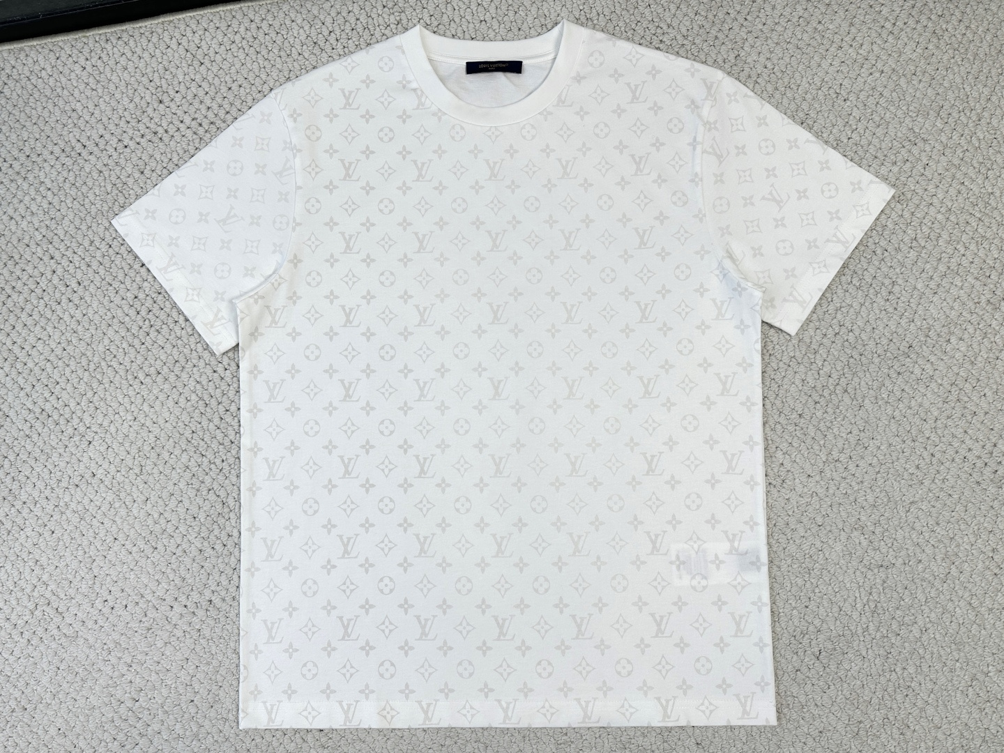 NO:914773,LV, Louis Vuitton, fluorescent printed T-shirt. Fabric: 15-count special woven compact cotton / 230G rib with 32-count 1×1 rib / 340G  Compact spinning technology effectively eliminates yarn hairiness, making the yarn body smooth and even, and the surface of the fabric presents a slightly matte high-end texture with excellent anti-pilling properties. The 15-count medium and low yarn count gives the fabric a natural crisp and skeletal feel, and the medium-high weight has both a drapey feel and a flexible frame.  , can naturally fit the body curve while maintaining a clear outline. The high-density ribbing on the neckline has a tight structure, solid texture, and excellent elasticity and resilience. It can firmly lock the collar and is not easy to loosen after long-term wear. It provides a smooth and stable base fabric for all-over printing.  This is the core technological highlight of the entire T-shirt. The all-over presbyopia pattern is printed on the fabric with a special photoluminescent slurry through screen printing. The luminous powder in the slurry shows different effects when exposed to ultraviolet light. Under normal light, the pattern appears as a same-color effect with consistent tones, low-key and restrained;  When placed in the dark and irradiated by an ultraviolet lamp, the stored energy is slowly released in the form of low light, making the Monogram pattern clearly show a unique fluorescent color. This process requires the printing paste to be evenly adhered to ensure a long-lasting and uniform reflective effect, and present a completely different visual appearance during the day and night.  Full-piece quilting is the key process to ensure the visual unity of the whole body pattern. Every piece from the body, sleeves to side seams must be strictly aligned with the printed pattern when cutting and sewing. The position and pattern direction of each piece need to be accurately calculated during the laying stage; single piece cutting is often used to ensure accuracy; during the sewing process, the seamstress needs to constantly adjust the fabric during sewing to ensure that the patterns at shoulder seams, side seams and other joints are seamlessly connected and the lines are consistent.,, louis  vuitton,louis vuitton,tees,t-shirt,19860909Lv,路易威登,荧火印花T恤 面料15支特纺紧密棉 / 230G 罗纹搭配32支1×1罗纹 / 340G 紧密纺技术有效消除纱线毛羽,使纱体光洁、条干均匀,面料表面呈现微雾面高级质感,抗起毛球性能优异.15支中低纱支赋予面料天然的挺括骨感,中高克重兼具垂坠感与柔韧骨架,能自然贴合身体曲线同时保持清晰轮廓.领口高密度罗纹结构紧密、质地扎实,具备极佳弹力与回弹性,能牢牢锁住领圈,经久穿着不易松弛,为通体印花提供了平整稳定的底布基础. 这是整件T恤最核心的工艺亮点.全副通体老花图案通过丝网印刷将含有特殊光致发光浆料印制于面料之上,浆料中的夜光粉在紫外光照射时呈现不同效果.正常光线下,图案呈现为色调一致的同色系效果,低调内敛；当置于黑暗中经紫外线灯照射时,储存的能量以微光形式缓慢释放,使Monogram图案清晰显现出独特的荧光色彩.该工艺要求印花浆料均匀附着,以确保反光效果持久均匀,白天与黑夜呈现截然不同的视觉面貌. 全件对花缝制是确保通体图案视觉统一的关键工艺.从大身、袖子到侧缝的每一片裁片,在裁剪和缝制时必须严格对齐印花图案.排料阶段需精确计算每片裁片的位置与图案走向；裁剪时多采用单片裁剪以保证精度；缝制过程中,缝纫工需在车缝时不断调整面料,确保肩缝、侧缝等拼接处的图案无缝连接、纹路贯通.,,louis vuitton,louis vuitton,tees，t-shirt,,Men's clothing
