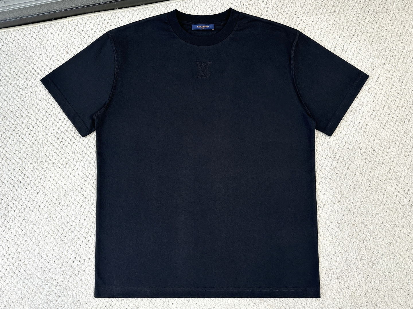 NO:956348,Lv Product Introduction 1V / Embossed T-shirt Fabric 15-count low-count cotton yarn / 230G Rib 32-count 1×1 rib / 340G This T-shirt uses cotton knitting to interpret the classic design, making it a good choice for layering daily looks. The embossed logo under the collar, the shoulder seams and the woven logo label on the back present an inside-out flip effect, highlighting unique creativity with dynamic strokes.  15-count low-count cotton yarn is selected, and the compact spinning process makes the fibers more condensed during the twisting process and reduces hairiness. The fabric has both a thick feel and a delicate surface. This fabric is stiff but not stiff, can bear subsequent embossing and is not easy to rebound. Utilizes high  The conflicting combination of heavy ribs and fine yarn fabrics achieves a thick yet delicate feel. Its function is that the neckline needs long-lasting tight elasticity. Thick ribs can resist lateral stretching and can be worn repeatedly without losing shape, which is the key to the durability of the T-shirt.  The LV logo indentation is made by pressing the mold at high temperature, and the fiber is heat-set in the process to form a permanent three-dimensional pattern. Then the computer embroidery machine performs dense stitch embroidery along the embossed edge, and the embroidery thread locks the embossed edge like a border. Its function is to emphasize the outline of the pattern with linear luster, and to strengthen the structural strength of the area to prevent the indentation from being damaged due to friction, so that the decorative part will last forever.  By overlapping the front and back pieces in reverse, the seams and overlay lines are neatly displayed on the outside of the shoulders, forming a regular chain-like decorative line. It breaks the monotony of the flat shoulders, uses changes in stitch thickness to modify the shoulder shape, and at the same time moves the visual center of gravity upward.,, louis vuitton, tees, t-shirt,19860909Lv 产品简介 1V / 压纹T恤 面料15支低支数棉纱 / 230G 罗纹32支1×1罗纹 / 340G 本款T恤以棉质针织演绎经典设计,成就日常造型的叠搭佳选.衣领下方压印标识,肩部缝线和背后的梭织标识标签呈现内外翻转效果,以活力笔触彰显别致创意. 选用15支低支数棉纱,经紧密纺工艺使纤维在加捻过程中更凝聚,降低毛羽,面料兼具厚实感与细腻表面.这种面料挺而不僵,能承载后续压纹而不易回弹.利用高克重罗纹与细纱面料的冲突组合,实现了既厚实又细腻的手感.其作用在于领口需要持久的紧致弹性,厚罗纹能抵抗横向拉伸,反复穿脱不走形,是T恤耐用性的关键. 以模具高温压烫出LV标识凹痕,纤维在此过程中被热定型,形成永久性立体纹路.随后电脑绣花机沿压纹边缘进行密针包边刺绣,绣线如镶边一般锁固压纹边缘.其作用一是以线性光泽强调图案轮廓,二是加强该区域的结构强度,避免压痕处因摩擦率先破损,使装饰部位历久弥新. 通过将前后衣片反向叠合,使得缝份和拷边线规整地展现在肩部外侧,形成规整的链条状装饰线.打破了平整肩部的单调,利用线迹粗细变化修饰肩型,同时让视觉重心上移.,,louis vuitton,tees，t-shirt,,Men's clothing
