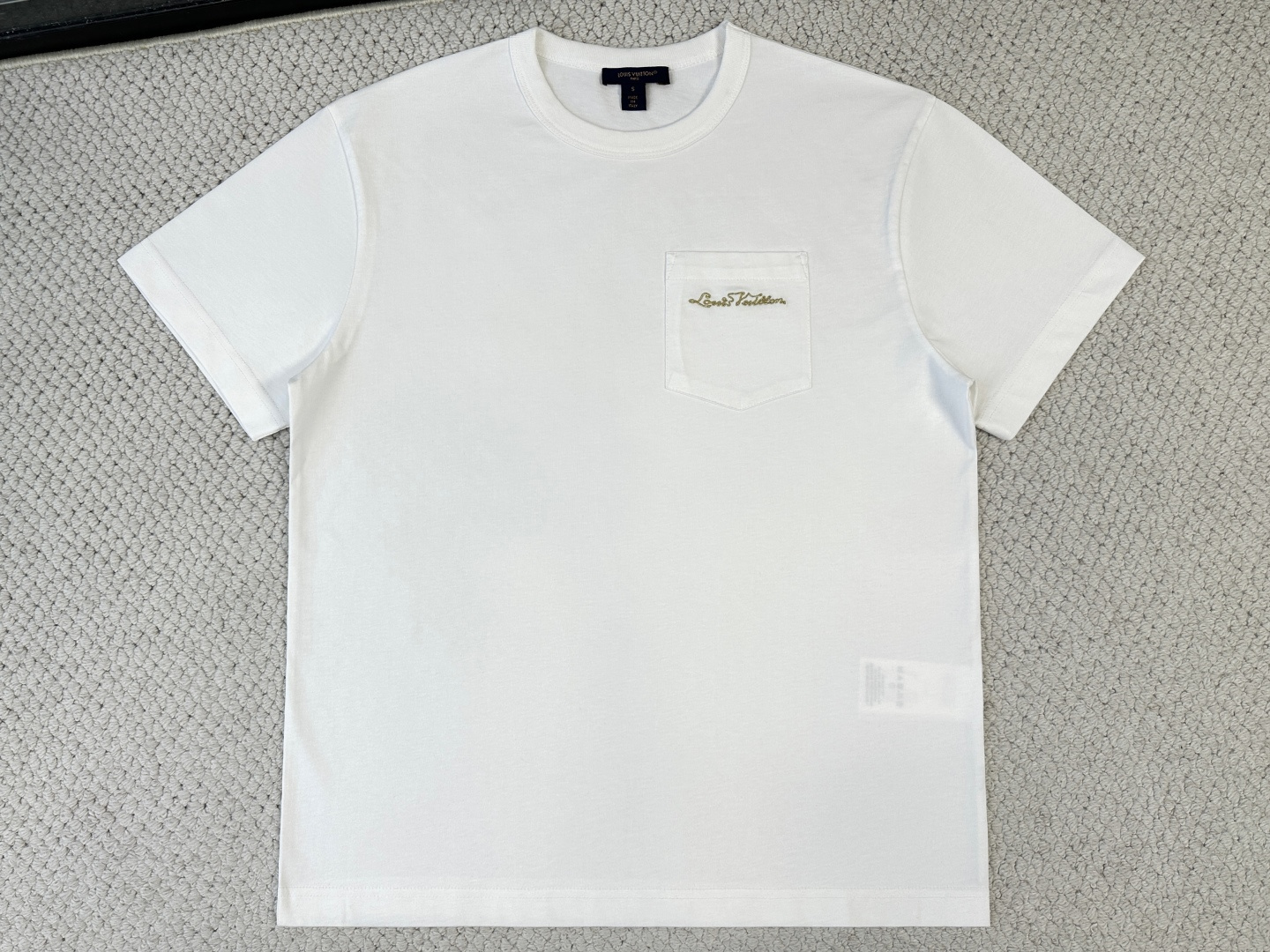 NO:956406,Lv. Product Introduction 1V / Logo Pocket T-shirt Fabric 15-count special woven compact cotton knitted fabric / 230G Rib 32-count high-density 1×1 rib / 340G This T-shirt is made of soft and breathable cotton knitted fabric, with a ribbed round neck for a straight fit. It is equipped with a sharp corner patch pocket on the chest, embellished with a metallic embroidered handwritten logo, which fashionably highlights the brand imprint.  It uses 15-count special-spun compact cotton knitted fabric. The weight of the fabric gives the fabric a rough natural texture and crisp bone support. The special-spun compact spinning technology makes the yarn hairiness minimal and even, and the cloth surface is smooth and tight, with both rough style and fine texture.  It has a greasy touch. The neckline is equipped with 32 high-density 1×1 ribs, and the ultra-high weight provides a structural support frame to ensure that the collar shape remains crisp after being worn and washed many times. The black and white base provides a pure canvas for the metal embroidery and three-dimensional pockets.  The sharp corner patch pocket on the chest is made of the same 15-count compact cotton fabric, and the bottom is gathered into a sharp angle, breaking the gentleness of traditional rounded corner pockets and injecting a neat and modern temperament. The edges are precision hemmed and hot-melted to ensure that the lines at the sharp corners are clean and sharp and will not turn up. The bag body is reinforced with high-density double-needle topstitching, forming a subtle layering and architectural outline with the body.  The top of the pocket is embellished with a metallic embroidered handwritten logo, using fine gold thread stitching technology. The stitches are closely arranged to form a smooth and continuous metallic luster surface. The gold thread is made of metalized polyester silk, which presents a liquid metal-like fluidity under the light. During embroidery, a hot-melt lining is inserted on the back of the pocket fabric to prevent the dense stitches from causing wrinkles in the fabric. The lining is melted after embroidery to ensure that the bag surface is flat and the sharp corners are not deformed by stress.,, louis vuitton, tees, t-shirt,19860909Lv. 产品简介 1V / 标识口袋T恤 面料15支特纺紧密棉针织面料 / 230G 罗纹32支高密1×1罗纹 / 340G 本款T恤取材柔软透气的棉质针织,为直身版型融入罗纹圆领.胸前配有尖角贴袋,其上点缀金属质感刺绣手写标识,时髦彰显品牌印记. 采用15支特纺紧密棉针织面料,面料克重赋予织物粗粝的天然肌理与挺括的骨感支撑.特纺紧密纺技术使纱线毛羽极少、条干均匀,布面光洁紧密,兼具粗犷风格与细腻触感.领口搭配32支高密1×1罗纹,超高克重提供结构性支撑框架,确保多次穿洗后领型仍保持挺括形态.黑色与白色基底为金属刺绣与立体口袋提供纯粹画布. 胸前尖角贴袋以同款15支紧密棉面料裁制,底部收拢成锐角,打破传统圆角口袋的温和感,注入利落摩登气质.边缘经精密折边与热熔定型处理,确保尖角处线条干净锐利、不翻翘.袋体通过高密度双针明线加固,与衣身形成微妙的层次感与建筑感轮廓. 口袋上方点缀金属质感刺绣手写标识,采用金线细挨针绣工艺,针脚紧密排列形成光滑连贯的金属光泽表面.金线由金属化涤纶丝制成,在光线下呈现液态金属般的流动感.刺绣时在口袋面料背面垫入热熔衬,防止密集针迹导致面料起皱,绣后衬料烫融确保袋面平整,尖角处不受力变形.,,louis vuitton,tees，t-shirt,,Men's clothing