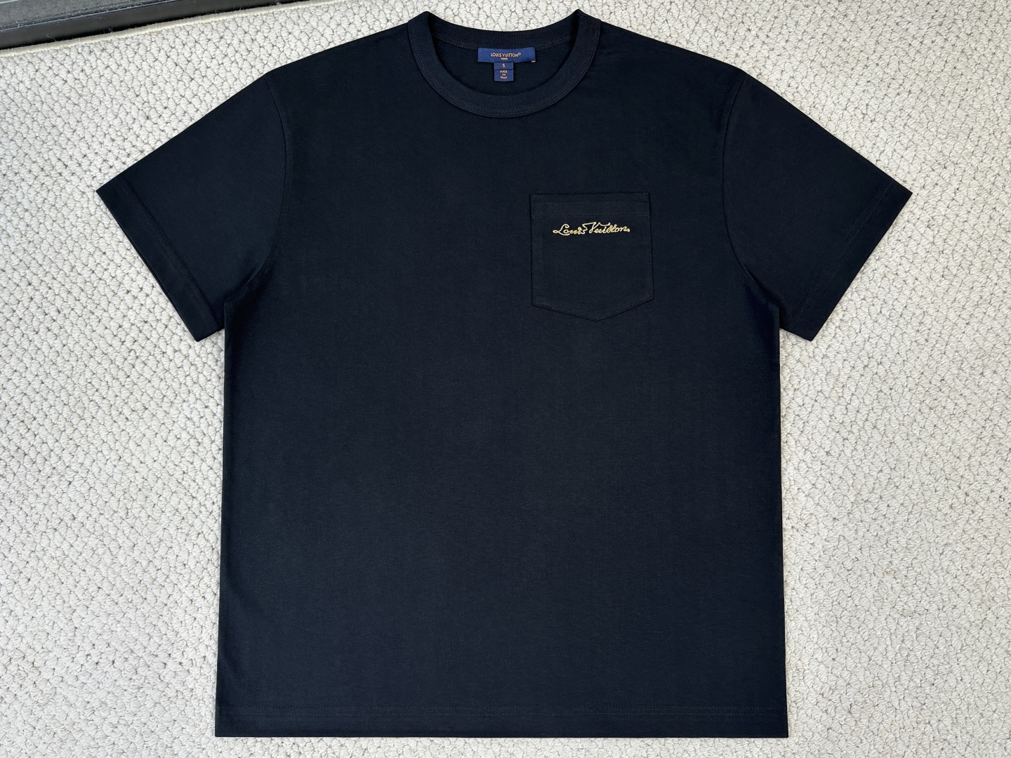 NO:956339,Lv Louis Vuitton Product Introduction 1V / Logo Pocket T-shirt Fabric 15-count special woven compact cotton knitted fabric / 230G Rib 32-count high-density 1×1 rib / 340G This T-shirt is made of soft and breathable cotton knitting, with a ribbed round neck for a straight fit. It is equipped with a sharp corner patch pocket on the chest, embellished with a metallic embroidered handwritten logo, which fashionably highlights the brand imprint.  It uses 15-count special-spun compact cotton knitted fabric. The weight of the fabric gives the fabric a rough natural texture and crisp bone support. The special-spun compact spinning technology makes the yarn hairiness minimal and even, and the cloth surface is smooth and tight, with both rough style and fine texture.  It has a greasy touch. The neckline is equipped with 32 high-density 1×1 ribs, and the ultra-high weight provides a structural support frame to ensure that the collar shape remains crisp after being worn and washed many times. The black and white base provides a pure canvas for the metal embroidery and three-dimensional pockets.  The sharp corner patch pocket on the chest is made of the same 15-count compact cotton fabric, and the bottom is gathered into a sharp angle, breaking the gentleness of traditional rounded corner pockets and injecting a neat and modern temperament. The edges are precision hemmed and hot-melted to ensure that the lines at the sharp corners are clean and sharp and will not turn up. The bag body is reinforced with high-density double-needle topstitching, forming a subtle layering and architectural outline with the body.  The top of the pocket is embellished with a metallic embroidered handwritten logo, using fine gold thread stitching technology. The stitches are closely arranged to form a smooth and continuous metallic luster surface. The gold thread is made of metalized polyester silk, which presents a liquid metal-like fluidity under the light. During embroidery, a hot-melt lining is inserted on the back of the pocket fabric to prevent the dense stitches from causing wrinkles in the fabric. The lining is melted after embroidery to ensure that the bag surface is flat and the sharp corners are not deformed by stress.,, louis vuitton, louis  vuitton,tees,t-shirt,19860909Lv 路易威登 产品简介 1V / 标识口袋T恤 面料15支特纺紧密棉针织面料 / 230G 罗纹32支高密1×1罗纹 / 340G 本款T恤取材柔软透气的棉质针织,为直身版型融入罗纹圆领.胸前配有尖角贴袋,其上点缀金属质感刺绣手写标识,时髦彰显品牌印记. 采用15支特纺紧密棉针织面料,面料克重赋予织物粗粝的天然肌理与挺括的骨感支撑.特纺紧密纺技术使纱线毛羽极少、条干均匀,布面光洁紧密,兼具粗犷风格与细腻触感.领口搭配32支高密1×1罗纹,超高克重提供结构性支撑框架,确保多次穿洗后领型仍保持挺括形态.黑色与白色基底为金属刺绣与立体口袋提供纯粹画布. 胸前尖角贴袋以同款15支紧密棉面料裁制,底部收拢成锐角,打破传统圆角口袋的温和感,注入利落摩登气质.边缘经精密折边与热熔定型处理,确保尖角处线条干净锐利、不翻翘.袋体通过高密度双针明线加固,与衣身形成微妙的层次感与建筑感轮廓. 口袋上方点缀金属质感刺绣手写标识,采用金线细挨针绣工艺,针脚紧密排列形成光滑连贯的金属光泽表面.金线由金属化涤纶丝制成,在光线下呈现液态金属般的流动感.刺绣时在口袋面料背面垫入热熔衬,防止密集针迹导致面料起皱,绣后衬料烫融确保袋面平整,尖角处不受力变形.,,louis vuitton,louis vuitton,tees，t-shirt,,Men's clothing