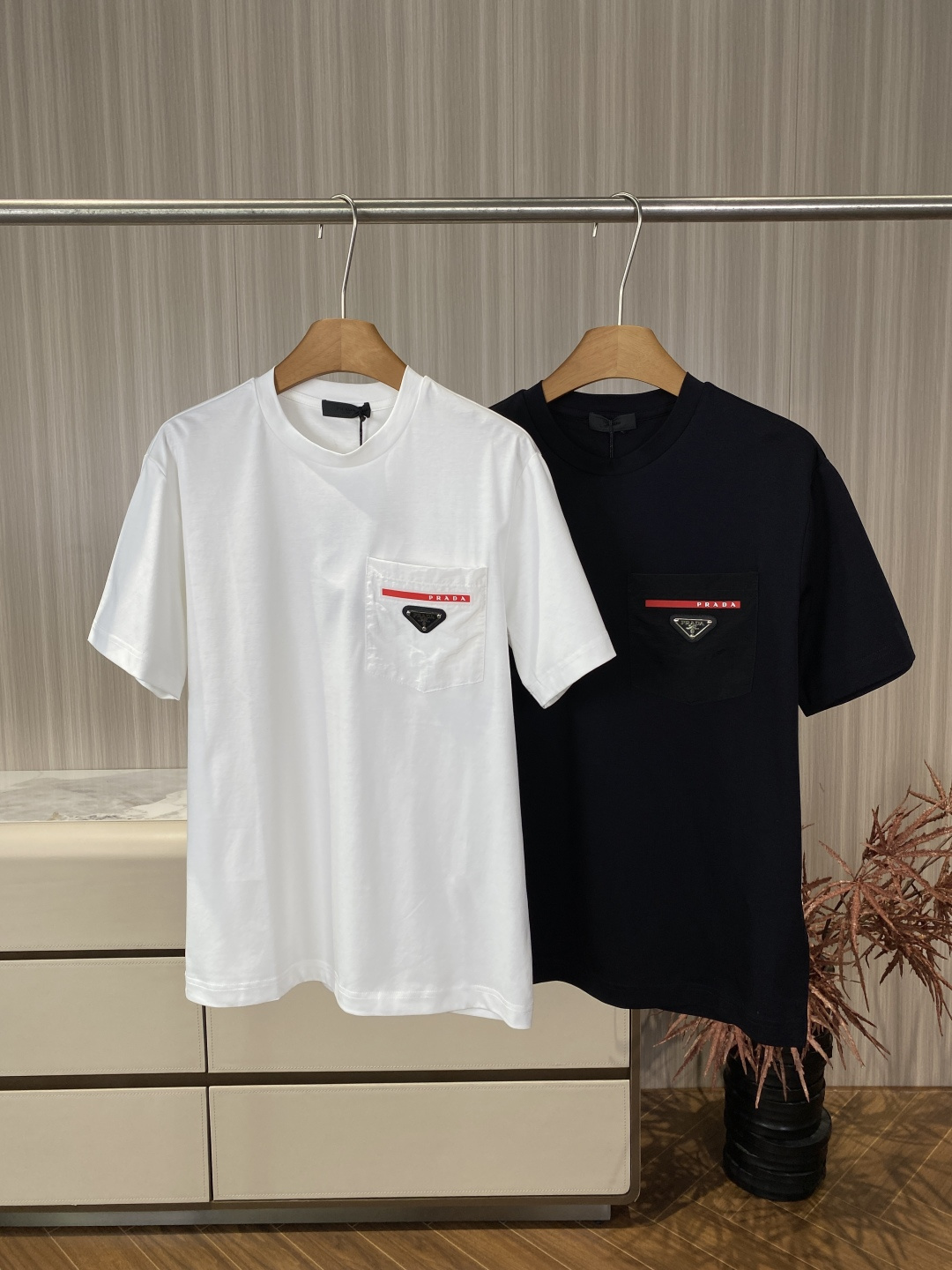 NO:835121,Prada Pocket Inverted Triangle Logo Short-Sleeved T-Shirt Prada's classic functional pocket short-sleeved T-shirt is designed with the iconic nylon pocket and inverted triangle logo combination as the core design. It is a representative item of the brand's 'luxury casual' style. 1. Iconic Nylon Pocket A nylon material patch pocket is used on the chest, which forms a material contrast with the pure cotton body, giving it both practicality and visual layering.  The top of the pocket is decorated with a PRADA webbing strip with white letters on a red background, echoing the inverted triangle metal logo below, which is a highly recognizable brand symbol. The inverted triangle metal logo is fixed with a black nylon base and silver rivets, and is engraved with the words 'PRADA MILANO'. It has a thick texture and exquisite details. 2. Minimalist silhouette and craftsmanship. Classic round neck + loose dropped shoulders. The silhouette is casual and not flattering, suitable for all body types.  The cuffs and hem are made of precision flat stitching, and the edges are smooth and not easily deformed after long-term wear. There is a black woven collar label sewn on the inside of the back collar, and it is paired with a black sling, which reflects the luxury-level details of the workmanship. The material is cotton, high-count combed cotton fabric. The fabric is soft and smooth, has good breathability and strong sweat absorption, and is suitable for spring and summer wear, and is skin-friendly and comfortable.  Care recommendations: Wash gently in cold water and dry on the inside. Avoid high-temperature ironing and drying to prevent fabric shrinkage and deformation. Wear functional casual style alone with overalls or sports shorts and dad shoes to create a street trend. Light business layering with a white turtleneck underneath and casual suits or windbreakers outside is suitable for commuting or semi-formal occasions. The universal basic style can be used as an inner layer of denim jackets and jackets, suitable for spring and autumn, to create a sense of layering.,, prada, shorts, jackets  & coats,jackets & coats,T-shirt,tees,t-shirt,business suit,19860909Prada 口袋倒三角标短袖T恤 Prada经典机能风口袋款短袖T恤,以标志性的尼龙口袋与倒三角Logo组合为核心设计,是品牌“奢华休闲”风格的代表单品. 1. 标志性尼龙口袋 胸口采用尼龙材质贴袋,与纯棉衣身形成材质对比,兼具实用性与视觉层次感. 口袋上方饰有红底白字PRADA织带条,与下方的倒三角金属标形成呼应,是辨识度极高的品牌符号. 倒三角金属标采用黑色尼龙底+银色铆钉固定,刻有“PRADA MILANO”字样,质感厚重,细节精致. 2. 极简廓形与工艺 经典圆领+宽松落肩剪裁,版型休闲不挑身材,适合各种体型穿着. 袖口与下摆采用精密平车缝线,边缘处理平整,久穿不易变形. 后领内侧缝有黑色织唛领标,搭配黑色吊绳,体现奢侈品级的细节做工. 材质棉,高支精梳棉面料. 触感与性能面料柔软顺滑,透气性佳,吸汗性强,适合春夏季节穿着,亲肤舒适. 护理建议冷水轻柔洗涤,反面晾晒,避免高温熨烫与烘干,防止面料缩水变形. 机能休闲风单穿搭配工装裤或运动短裤,脚踩老爹鞋,打造街头潮流感. 轻商务叠穿内搭白色高领打底,外穿休闲西装或风衣,适合通勤或半正式场合. 万能基础款可作为牛仔外套、夹克的内搭,适配春秋季节,营造层次感.,,prada,shorts,jackets & coats,jackets & coats,T-shirt,tees，t-shirt,business suit,,Women's clothing
