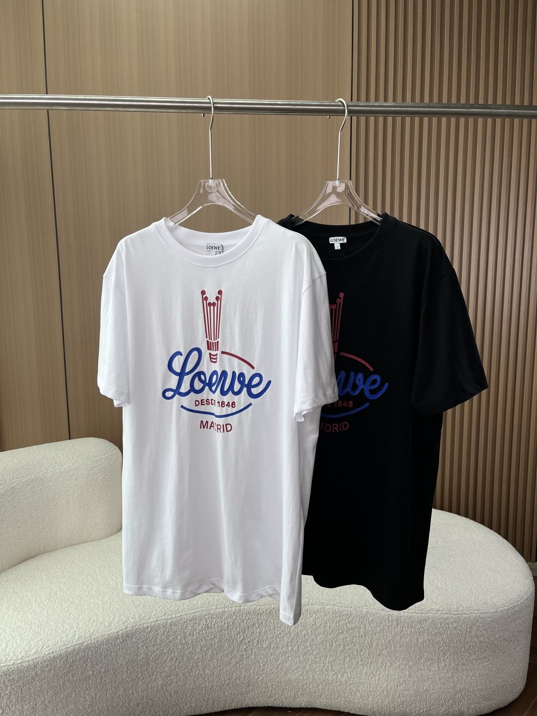 NO:832060,(Original customization) LOEWE retro crown printed short-sleeved T-shirt!  This is the retro American-style core item of LOEWE's 2026 spring and summer series. It is inspired by the brand's classic crown LOGO from the 1940s, with both retro texture and modern relaxation. 1. Style design 1. Core design elements Front printing The red retro crown pattern at the top restores the brand's classic LOGO from the 1940s, which is an iconic symbol of the brand's history; the LOEWE brand name in blue handwritten American retro cursive script in the middle is highly recognizable; the red small letters below  DESDE 1846 MADRID marks the year of the brand's founding (1846) and the place of birth (Madrid, Spain), strengthening the traceability of the brand. The back details include the LOEWE classic Anagram (four-way interweaving) logo embroidered under the back collar. The embroidery in the same color is low-key and unobtrusive. It is a detailed design that 'knows the brand at a glance'.  The silhouette adopts a loose, dropped-shoulder wide-cut, and is the same style for men and women. It is highly tolerant to all body shapes (skinny, slightly fat, and strong), hides the body, and creates an unintentional sense of relaxation. It is the most popular 'Oversize' style at the moment. The color options include pure white/pure black. White is refreshing and clean, suitable for spring and summer, creating an American casual and Japanese relaxed feel; black is cool and versatile, suitable for all seasons, creating a street and light business style.  2. Process details: The printing adopts environmentally friendly offset printing technology, the pattern is clear and full, there is no smearing on the edges, the touch is soft and not hard, and it will not crack or fall off after long-term washing. The white woven collar label, LOEWE letters are clearly printed, and are exclusive brand fonts. The collar label stitching is neat, and the inner folding hides the wiring.  , the tag is off-white, printed with the brand name intertwined with the Anagram logo, and the tag rope is a black double-stranded cotton rope. The Anagram embroidery on the back is neat, the stitches are fine, the pattern is symmetrical, and the stitches of the same color are integrated with the fabric, without jumpers or missing stitches. Thread routing: double-strand threading throughout the body, the collar, cuffs, and hem ribbed edges are full of elasticity, and will not loosen or deform after long-term wear. 2. Fabric 1. Fabric composition  According to the washing label, the fabric is a cotton-polyester blend with the following specific ingredients: Main fabric 35 cotton + 65 polyester fiber Rib part (collar/cuffs/hem) 35 cotton + 65 polyester fiber Embroidery part cotton (embroidery thread) Accessories part polyester fiber 2. Fabric advantages  Cotton-polyester blend combines the skin-friendly breathability of cotton with the anti-wrinkle and non-deformation resistance of polyester fiber. It solves the pain points of pure cotton T-shirts that are prone to wrinkles and shrinkage, and remains crisp and stylish after long-term wear. The heavyweight fabric has a thick texture, a full drape, and is not close-fitting to the body, creating a lazy and relaxed look.  Excellent breathability, no stuffiness when worn in spring and summer, can be worn in all seasons.,,loewe,loewe,versace,T-shirt,tees,t-shirt,19860909（原版定制） LOEWE（罗意威）复古皇冠印花短袖T恤！ 这是LOEWE 2026春夏系列的复古美式风核心单品,以品牌1940年代经典皇冠LOGO为设计灵感,兼具复古质感与现代松弛感 一、款式设计 1. 核心设计元素 正面印花 顶部红色复古皇冠图案还原品牌1940年代的经典LOGO,是品牌历史感的标志性符号； 中间蓝色手写体LOEWE品牌名美式复古花体字,辨识度拉满； 下方红色小字 DESDE 1846 MADRID标注品牌创立年份（1846年）与发源地（西班牙马德里）,强化品牌溯源. 后背细节后领下方绣有LOEWE经典Anagram（四向交织）logo,同色系刺绣低调不张扬,是“懂行一眼识品牌”的细节设计. 版型剪裁采用宽松落肩阔版剪裁,男女同款,对所有身形（瘦、微胖、壮硕）都有极强包容性,藏肉显瘦,打造不刻意的松弛感,是当下最火的“Oversize”穿搭版型. 配色选择提供纯白/纯黑两色 白色清爽干净,适配春夏,打造美式休闲、日系松弛感； 黑色酷飒百搭,适配全季节,打造街头、轻商务风格. 2. 工艺细节 印花采用环保胶印工艺,图案清晰饱满,边缘无晕染,触感柔软不发硬,久洗不开裂、不脱落,白色织唛领标,LOEWE字母印刷清晰,为品牌专属字体,领标缝线工整,内折隐藏走线 ,吊牌为米白色吊牌,印有品牌名与Anagram交织logo,吊牌绳为黑色双股棉绳.后背Anagram刺绣工整,针脚细密,图案对称,同色系线迹与面料融为一体,无跳线、漏针 走线 全身双股走线,领口、袖口、下摆罗纹收边弹性十足,久穿不松垮、不变形 二、面料 1. 面料成分 根据洗标标注,面料为棉涤混纺,具体成分如下 主体面料35棉 + 65聚酯纤维 罗纹部分（领口/袖口/下摆）35棉 + 65聚酯纤维 刺绣部分棉（绣线） 辅料部分聚酯纤维 2. 面料优势 棉涤混纺兼顾了棉的亲肤透气与聚酯纤维的抗皱、不易变形,解决了纯棉T恤易皱、易缩水的痛点,久穿依然挺括有型； 重磅面料质感厚实,垂坠感拉满,上身不贴身,打造慵懒松弛的穿搭效果； 透气性优秀,春夏穿着无闷热感,四季可穿.,,loewe,loewe,versace,T-shirt,tees，t-shirt,,Women's clothing