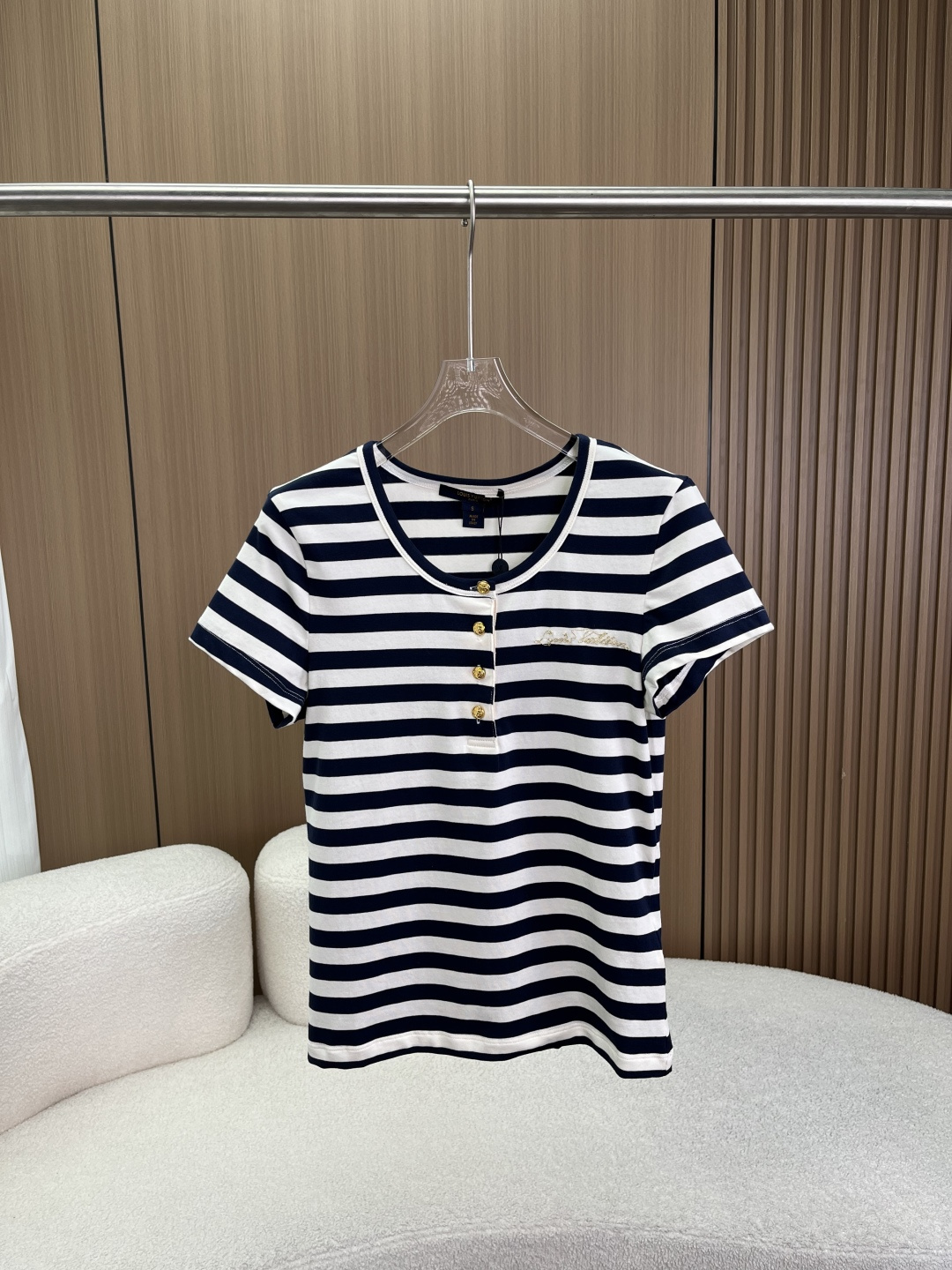 NO:874983,LV Louis Vuitton's new blue and white striped Henley collar short knitted T-shirt!  Made of 190g high-density knitted striped fabric, with classic blue and white contrasting horizontal stripes as the visual core, and precise yarn-dyed stripe weaving technology to achieve clear boundaries and even arrangement between strips. The fabric has both a delicate skin-friendly touch and excellent drape shaping ability, adapting to the needs of slim and short tailoring.  The ready-made garment adopts a U-shaped Henry collar placket design, with 4 custom metal snap buttons embellished with vintage distressed gold craftsmanship, which is both practical and decorative. The collar uses contrasting edge rolling technology to enhance the stiffness and durability of the collar, while echoing the contrasting aesthetics of the stripes on the body. The left chest uses flat embroidery to present the brand's handwritten logo, using high-density flat stitch embroidery. The stitches are dense and full, with a strong three-dimensional effect, forming a distinct visual hierarchy on the striped base fabric.  The sleeves are cut with right-shoulder short sleeves, and the cuffs are processed with double-strand seaming technology to ensure neat routing and washability; the body is cut with a slim waist, and the short length design optimizes body proportions and creates a retro hot girl style. The moderate weight of 190g combines breathability and perspiration for spring and summer wear with wrinkle resistance for daily wear. The yarn-dyed stripe process avoids the risk of the printed stripes falling off, and the flat embroidery logo uses traditional embroidery technology to enhance the craftsmanship value and sense of luxury of the item., louis  vuitton,louis vuitton,T-shirt,tees,t-shirt,19860909LV路易威登新款蓝白条纹亨利领短款针织T恤！ 采用190g高密针织条纹面布打造,以经典蓝白撞色横条纹为视觉核心,通过精准的色织条纹织造工艺实现条带间的清晰分界与均匀排布,布面兼具细腻亲肤触感与优异的悬垂塑形力,适配修身短款剪裁的版型需求 成衣采用U型亨利领门襟设计,门襟处搭载4颗定制金属四合扣,以复古做旧金工艺点缀,兼具开合实用性与装饰性；领圈采用撞色包边滚条工艺,强化领型挺括度与耐穿性,同时呼应衣身条纹的撞色美学.左胸位置以平绣工艺呈现品牌手写体标识,采用高密平针绣法,针脚细密饱满、立体感强,在条纹底布上形成鲜明的视觉层次 袖身采用正肩短袖剪裁,袖口以双股线锁边工艺处理,保证走线工整与耐洗性；衣身采用修身收腰剪裁,短款衣长设计优化身材比例,打造复古辣妹风格.190g适中克重兼顾春夏穿着的透气排汗性与日常穿着的抗皱性,色织条纹工艺避免了印花条纹的脱落风险,平绣标识则以传统刺绣工艺提升单品的工艺价值与高级感,,louis vuitton,louis vuitton,T-shirt,tees，t-shirt,,Women's clothing