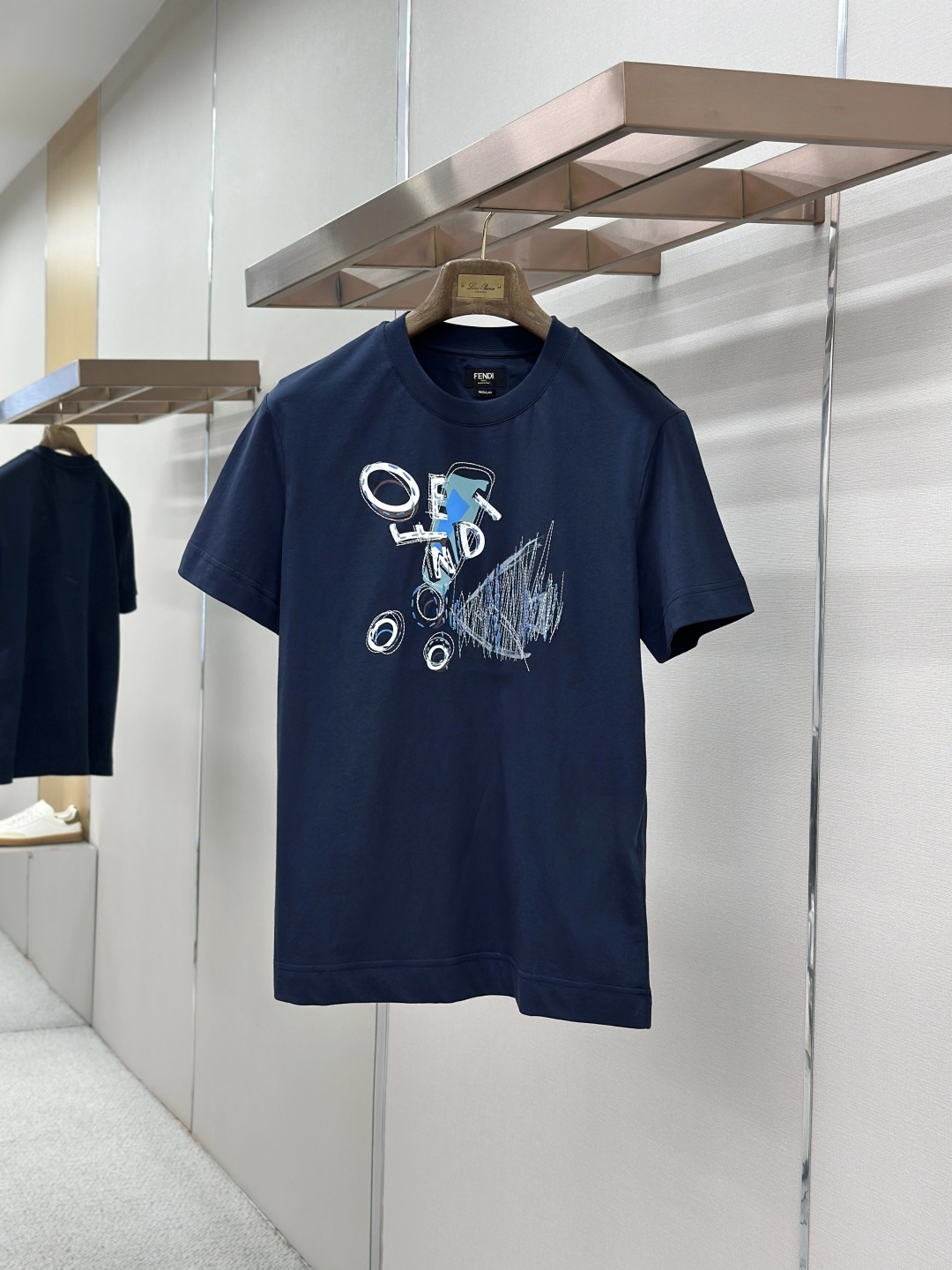 NO:857352,Fendi Fendi-Goldfish pattern cotton T-shirt. Fabric: 24-count double-sided knitted spun yarn 250G. Rib: 24-count 1×1 rib 200G. Standard fit short-sleeved round neck T-shirt, made of dark blue cotton. Decorated with graphic print and yarn topstitch details on the front. Rib knit crew neck.  Count 24 is a mid-to-high fine yarn count, the yarn is thin, and the fabric surface is delicate and smooth. The double-sided knitted structure has a front-facing appearance on both sides. The medium-thick weight gives the fabric a soft and crisp feel; it has the unique fluffy feel of double-sided knitting without losing drape support, and the upper body is not soft. High color fastness reactive dye dyeing ensures that the dark background color remains full after multiple washes, providing a smooth and stable base fabric for the triple-technical composite of the goldfish pattern.  The front goldfish pattern achieves the symbiosis of flat color and three-dimensional texture through the precise cooperation of environmentally friendly elastic glue, dot printing and printed multi-color rice embroidery. The environmentally friendly elastic glue is a water-based polyurethane-based slurry, and the film has a very high elongation at break after curing.  It can be stretched synchronously with the extension of the fabric and will not crack after repeated washing. Dot printing transfers the slurry in the form of micron-level dots through a high-mesh screen plate to achieve photo-level grayscale gradient and half-tone effects, avoiding the color block faults common in glue printing.  The printed multi-color rice grain embroidery is a decorative embroidery stitch. Each needle is short and independent, shaped like a rice grain, forming a dot-like texture on the cloth surface; the embroidery machine accurately places each needle on a specific area of the printed pattern, using mercerized cotton embroidery thread with a soft luster, and selectively embellishing the details of the goldfish pattern. Sizes XS-L,, fendi, fendi, T-shirt, tees, t-shirt,19860909Fendi芬迪-金鱼图案棉质T恤 面料24支双面针织细纱 250G 罗纹24支1×1罗纹 200G 标准版型短袖圆领T恤,深蓝色纯棉材质.正面饰有图形印花和纱线明线细节.罗纹针织圆领. 24支属于中高细纱支,纱线较细,布面细腻光滑.双面针织结构由具有两面皆为正面的外观.中厚克重赋予面料柔软的挺括感；既有双面针织特有的蓬松手感,又不失垂坠支撑力,上身不软塌.高色牢度的反应性染料染色确保了深色底色在多次洗涤后依然饱满,为金鱼图案的三重工艺复合提供了平整稳定的底布基础. 正面金鱼图案通过环保弹力胶浆、网点印花与对印花多色米粒绣的精密配合,实现平面色彩与立体肌理的共生.环保弹力胶浆为水性聚氨酯基浆料,固化后薄膜断裂延伸率极高,能跟随面料延展同步拉伸,反复洗涤后不龟裂.网点印花通过高目数网版将浆料以微米级点阵形式转移,实现照片级的灰度渐变和半色调效果,避免了胶浆印花常见的色块断层. 对印花多色米粒绣为装饰性刺绣针法,每针短小独立、形似米粒,在布面形成点状肌理；绣花机将每一针精确落在印花图案的特定区域,选用丝光棉绣线具有柔和光泽,选择性地点缀金鱼图案细节部位. 尺码XS-L,,fendi,fendi,T-shirt,tees，t-shirt,,Men's clothing