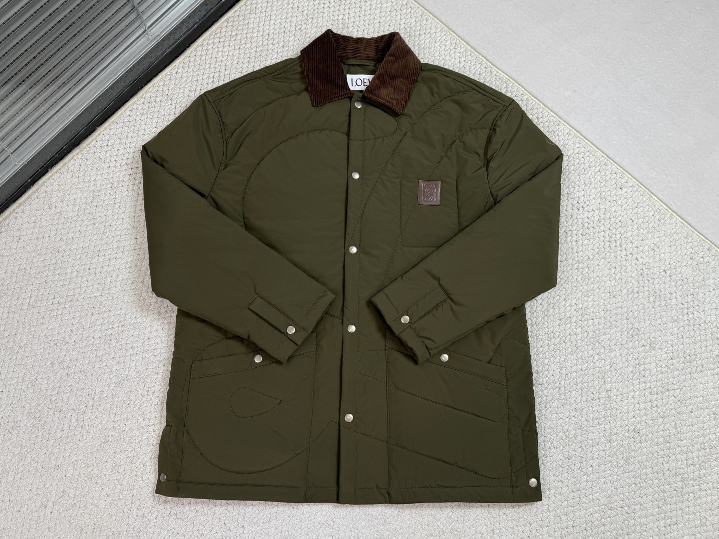 NO:610185,Loewe, Loewe - quilted technical fabric padded jacket shirt. The main body is made of high-performance polyester fiber, which is lightweight and warm through a three-layer composite structure. The ultra-thin padding is combined with double-sided three-dimensional quilting technology to accurately outline the brand logo with a density of 12 inches, which not only effectively locks the filling to avoid displacement, but also achieves temperature regulation through the air barrier. The quilting depth is precisely calculated to produce subtle three-dimensional light and shadow changes during dynamic wearing.  The full polyester fiber lining has been treated with antistatic treatment and is perfectly wrapped with three-dimensional tailoring to wrap the internal structure. All seams are hemmed to achieve a friction-free wearing experience. The lining and top fabric are connected through a gapped three-dimensional suspension system to ensure that the inner and outer layers move independently during activities.  The collar is made of corduroy embossed suede, and parallel grooves are pressed on the suede surface through a Celsius high-temperature mold. This treatment not only retains the soft touch of the suede, but also adds a retro texture, forming a dramatic dialogue between futuristic and nostalgic aesthetics with the main technical fabric. Invisible inlay technology is used at all seams to ensure the smoothness of the transition between different materials.  The front placket adopts reinforced snap-button hardware, the cuffs have an innovative adjustable structure, and the side slits are equipped with hidden snap-buttons; the internal leather patch pockets are made of calfskin and laser-engraved, and the edges are waterproofed.19860909罗意威,Loewe-夹棉科技面料衬垫外套衬衫 主体采用高性能聚酯纤维,通过三层复合结构实现轻量化保暖.超薄衬垫与双面立体綗缝工艺结合,以寸12针的密度精准勾勒品牌标识,既有效锁定填充物避免移位,又通过空气隔层实现温度调节功能.绗缝深度经过精密计算,在动态穿着时产生微妙的立体光影变化. 全聚酯纤维衬里经过抗静电处理,以立体剪裁完美包裹内部结构,所有缝份采用卷边工艺处理,实现无摩擦穿着体验.里布与面布通过间隙的立体悬挂系统连接,确保活动时内外层独立运动. 衣领采用灯芯绒压纹绒面革,通过摄氏高温模具在绒面表面压制出平行沟槽.这种处理既保留绒面革的柔软触感,又增添复古纹理,与主体科技面料形成未来感与怀旧美学的戏剧性对话.所有接缝处采用隐形内接技术,确保不同材质间的过渡平整度. 前襟采用强化按扣五金配件,袖口创新可调节结构,侧开衩配置隐藏式按扣；内部皮革贴袋选用小牛皮经激光雕刻成型,边缘进行防水封边处理.,,loewe,loewe,jackets & coats,shirts,cowhide,alexander wang,Men's clothing