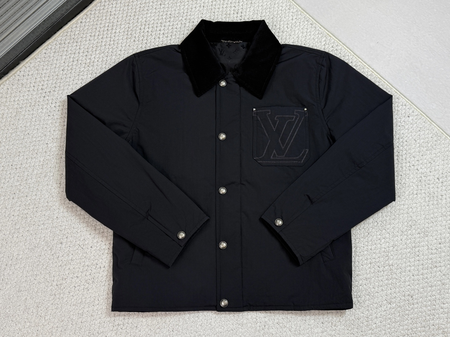 NO:645427,Lv Nylon Blend Work Jacket This work jacket is equipped with a sophisticated corduroy collar, suitable for all seasons. The water-repellent nylon blend body and quilted lining are both functional and warm. On top of the simple silhouette, the embroidery on the chest pocket and the leather patch on the back are the finishing touches. The zipper front is equipped with snap buttons, which is practical and elegant.  The body of the garment is made of a blended structure of 61 nylon and 39 cotton. The nylon gives the fabric water-repellent and abrasion resistance, while the cotton ensures skin-friendly breathability. The lining is a nylon quilted structure with built-in polyester fiber filler, which locks the air layer through precision stitching to achieve the dual functions of lightweight warmth and filling fixation.  The corduroy collar has a unique longitudinal velvet stripe texture that contrasts with the main functional fabric. The chest pocket logo uses a needle embroidery process, using dense stitches of 18 stitches per inch to outline the pattern to ensure the durability of the logo in workwear scenes. The laser-cut leather patch on the back uses luxurious materials to complete the closed loop of the brand narrative.  The innovative zipper and snap button dual structure hidden zipper ensures absolute wind resistance, and only the orderly beauty of the snap button is exposed on the outside. Both the cuffs and side pockets use military-grade snap components to achieve quick adjustment and safe storage functions.,,louis vuitton,jackets & coats,alexander wang19860909Lv 锦纶混纺工装夹克 这款工装夹克配备考究灯芯绒衣领,四季皆宜；防泼水锦纶混纺衣身与绗缝内里兼顾功能与保暖.简约廓形之上,胸袋的刺绣与背面的皮革贴饰成为点睛之笔.拉链前襟搭配按扣,实用而优雅. 衣身采用61锦纶与39棉的混纺结构,锦纶赋予面料防泼水与耐磨特性,棉质则保障亲肤透气性.内衬为锦纶绗缝结构,内置聚酯纤维填充物,通过精密线迹锁定空气层,实现轻量保暖与填充物固定双重功能. 灯芯绒衣领以独特的纵向绒条纹理,与主体功能面料形成质感对比.胸袋标识采用挨针刺绣工艺,通过每英寸18针的密实线迹勾勒图案,确保标识在工装场景下的耐久性.背面激光切割皮革贴饰则以奢华材质完成品牌叙事的闭环. 创新拉链与按扣双重结构隐藏式拉链保障绝对防风性,外表仅显露按扣的秩序美感.袖口与侧袋均采用军用级按扣组件,实现快速调节与安全储物功能.,,louis vuitton,jackets & coats,alexander wang,Men's clothing