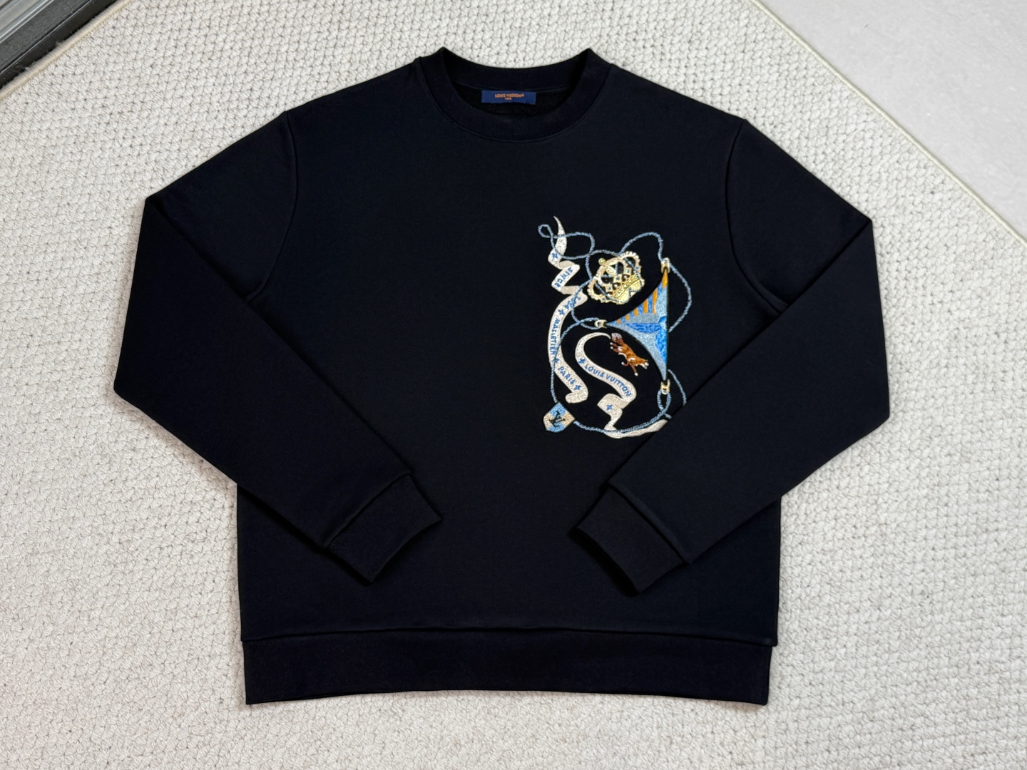 NO:712308,Lv-Product Introduction 1V / Embroidered long-sleeved sweater Fabric 26-count double yarn, bottom is 10-count yarn / 380G Rib 21-count 1×1 rib / 470G  This classic long-sleeved sweater has been sublimated with this season's emblem pattern. Its bright colors and noble symbols containing brand elements are exquisitely embroidered on the chest with bright silk threads. This design injects unique new ideas into the classic silhouette, adding a festive atmosphere and refined style to the casual look.  The double yarns on the top layer ensure that the cloth surface is delicate and smooth, and the bottom terry is precision brushed to form a thick suede surface, achieving the ultimate balance between the clean lines on the outside and the cloudy feel on the inside. The cuffs and hem are equipped with 21 super-heavy ribs of 470g, and the high-density weaving of 12 needles per inch increases the resilience by 60, completely eliminating sagging and deformation after wearing.  The overall pattern uses Zhangzi patch embroidery + multi-needle composite technology to first make a three-dimensional badge base with a thickness of independent embroidery pieces, then use stitch embroidery to outline the outline lines, and fill the color blocks with tatami embroidery. In the process, 17 colors of gold and silver embroidery threads are incorporated, and special twisting technology is used to interweave metal wires and cotton threads to produce a gradient streamer effect under the light. The overall embroidery surface presents a museum badge-level relief texture.  The loose but not bloated tailoring is designed with three-dimensional shoulders and a slight waistline to retain the street feel while optimizing the body proportions. All seams are overlocked and trimmed, and the internal seams are smooth and smooth. Double-layer reinforced sewing is performed on stress points such as collars and cuffs to ensure the structural stability of heavy embroidery and heavy fabrics.,, louis vuitton,19860909Lv-产品简介 1V / 刺绣长袖针织衫 面料26支双纱,底是10支纱 / 380G 罗纹21支1×1罗纹 / 470G 这款经典长袖针织衫,因本季的徽章图案而格调升华.其鲜亮色彩与蕴含品牌元素的尊贵符号,以莹亮丝线精美刺绣于胸前.这一设计为经典廓形注入别致新意,为休闲造型增添了节日氛围与精致格调. 面层双纱确保布面细腻光洁,底层毛圈经精密刷绒形成厚绒面,实现外部利落线条与内部云绒触感的极致平衡.袖口及下摆搭配21支470g超重磅罗纹,其每英寸12针的高密编织使回弹力提升60,彻底杜绝穿着后的松弛变形. 整体图案运用章仔贴布绣+多针法复合工艺先以独立绣片制成厚度的立体徽章基座,再运用挨针绣勾勒轮廓线条,并以榻榻米绣填充色块.过程中融入17色金银绣线,通过特殊捻线技术使金属丝与棉线交织,在灯光下产生渐变流光效果,绣面整体呈现博物馆徽章级的浮雕质感. 宽松而不臃肿的剪裁通过立体肩部与微收腰线设计,在保留街头感的同时优化身体比例.所有接缝采用锁边结合切边,内部缝份平整无感.领口、袖口等应力点位进行双层加固车缝,确保重工刺绣与厚重面料的结构稳定性.,,louis vuitton,,Men's clothing