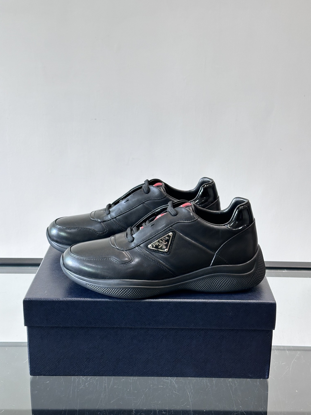 Prada黑色皮革运动鞋是品牌经典复古系列中的代表作，采用光滑小牛皮材质，搭配厚实橡胶大底和标志性的三角