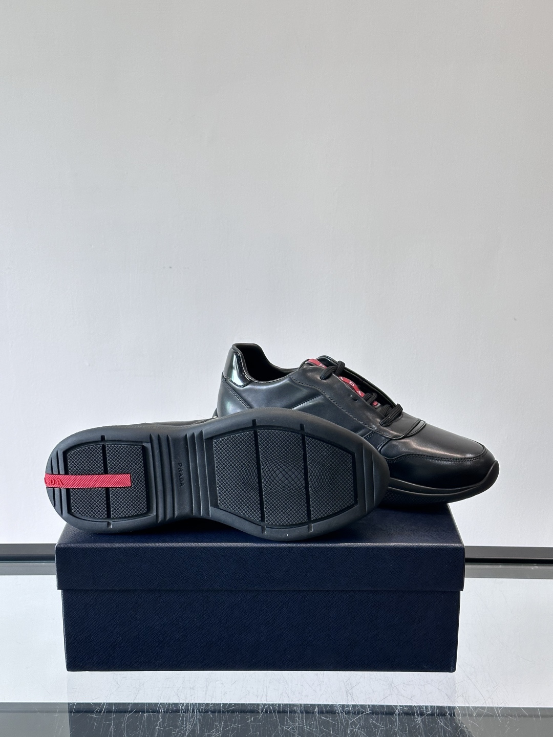 Prada黑色皮革运动鞋是品牌经典复古系列中的代表作，采用光滑小牛皮材质，搭配厚实橡胶大底和标志性的三角