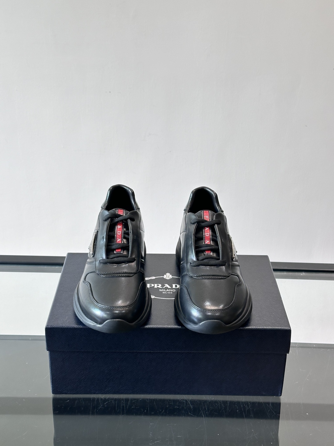 Prada黑色皮革运动鞋是品牌经典复古系列中的代表作，采用光滑小牛皮材质，搭配厚实橡胶大底和标志性的三角