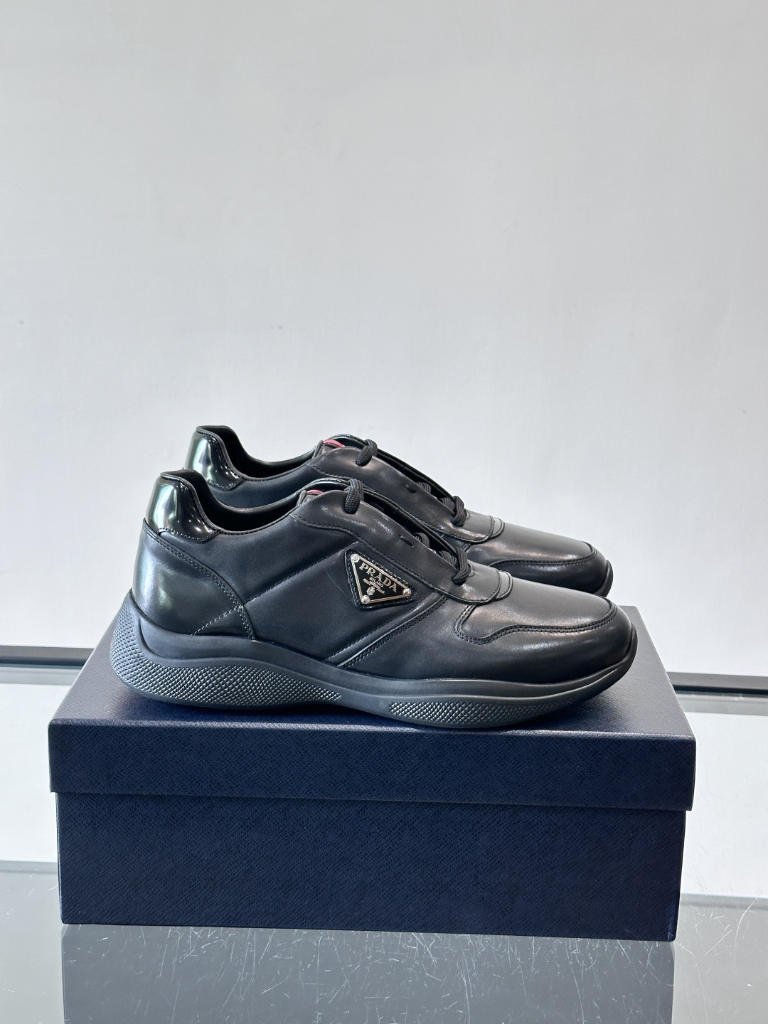 Prada黑色皮革运动鞋是品牌经典复古系列中的代表作，采用光滑小牛皮材质，搭配厚实橡胶大底和标志性的三角